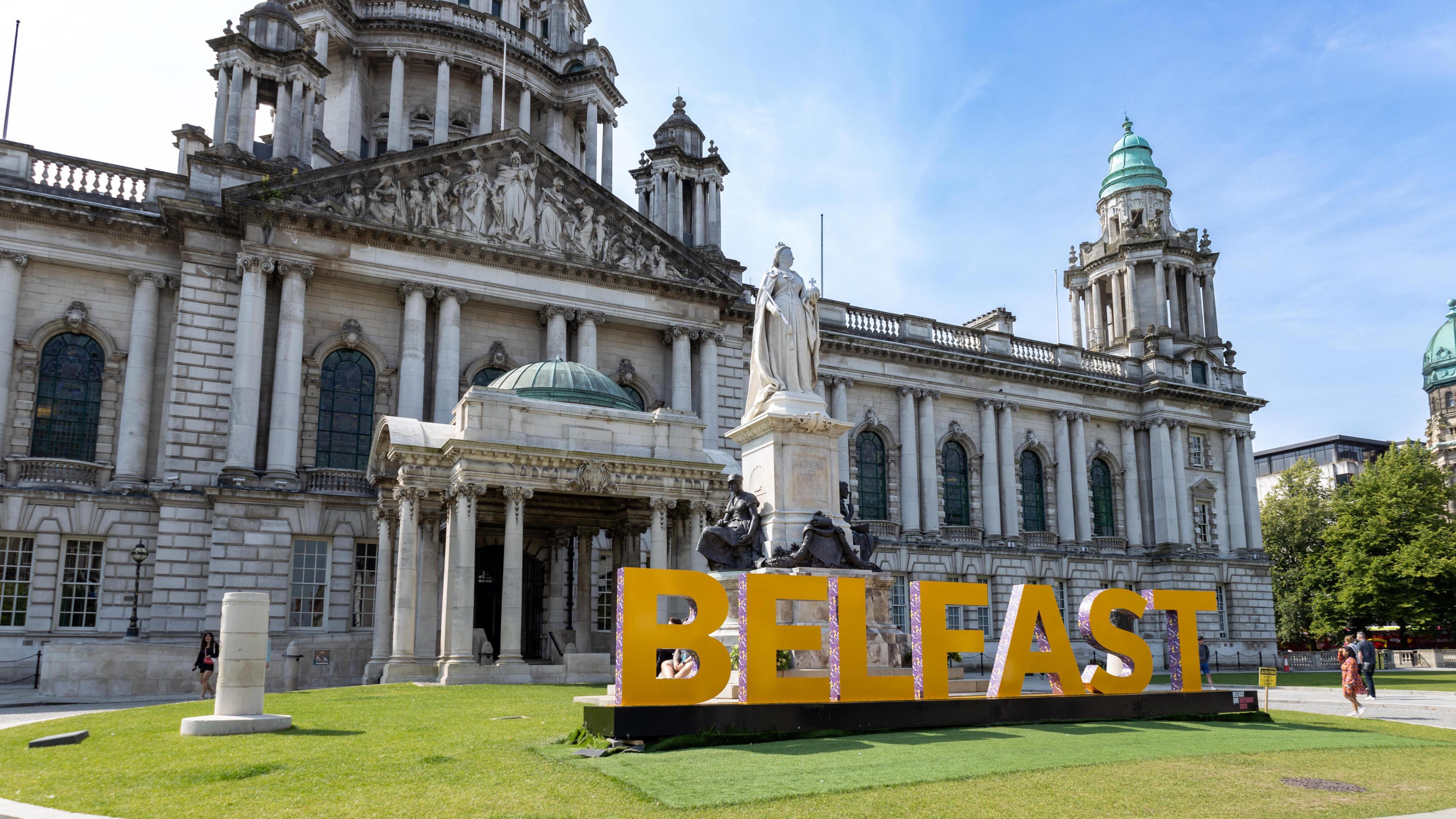 Belfast City Hall is a large white building with pillars and designs. There's a large statue of Queen Victoria at the front. Belfast in large letters are displayed on grass at the front of the building.