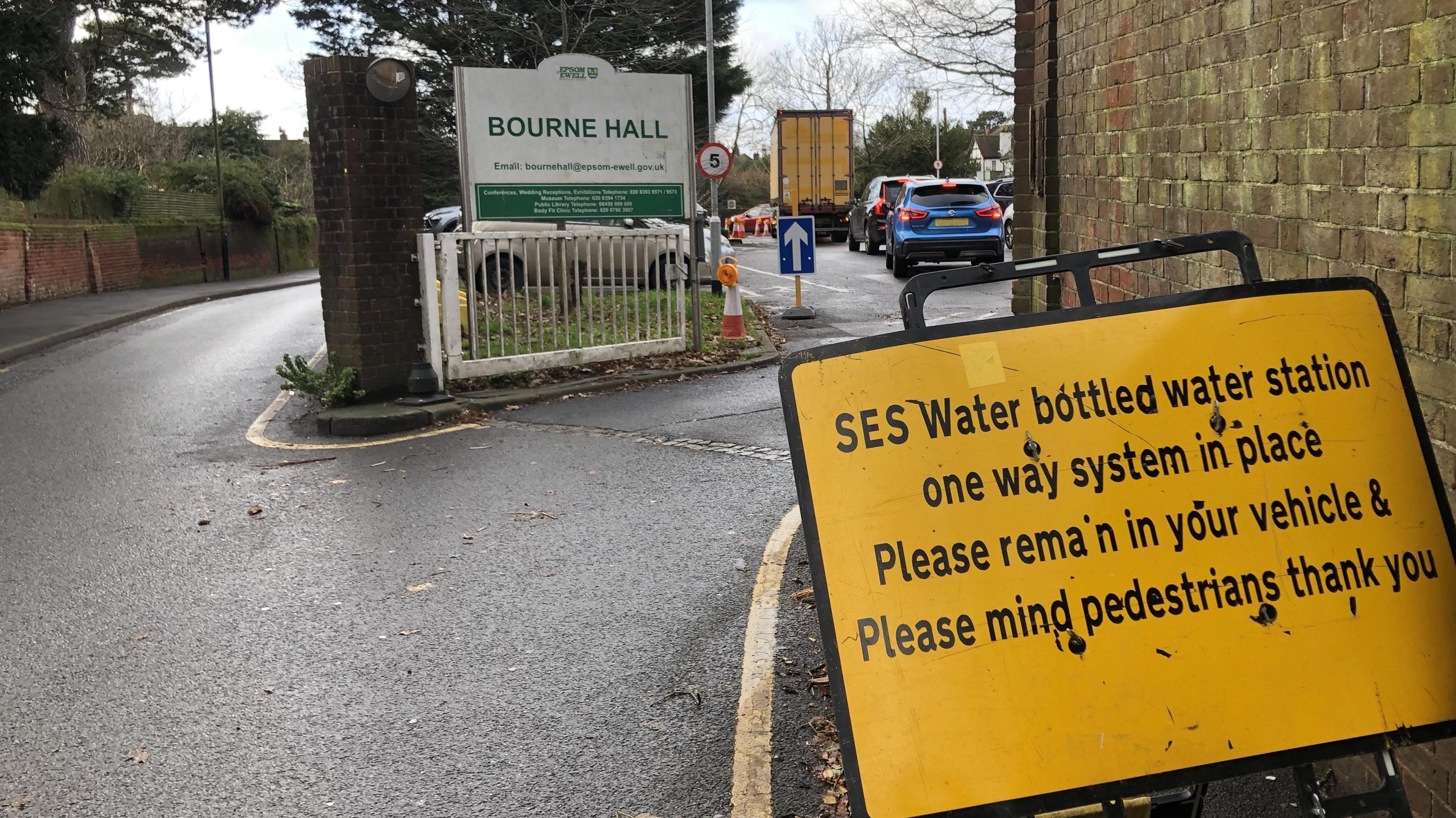 A yellow sign by SES water explaining the bottled water one way system sits by a wall, the entrance to Bourne Hall car park can be seen. In the distance, cars are queuing up for bottled water.