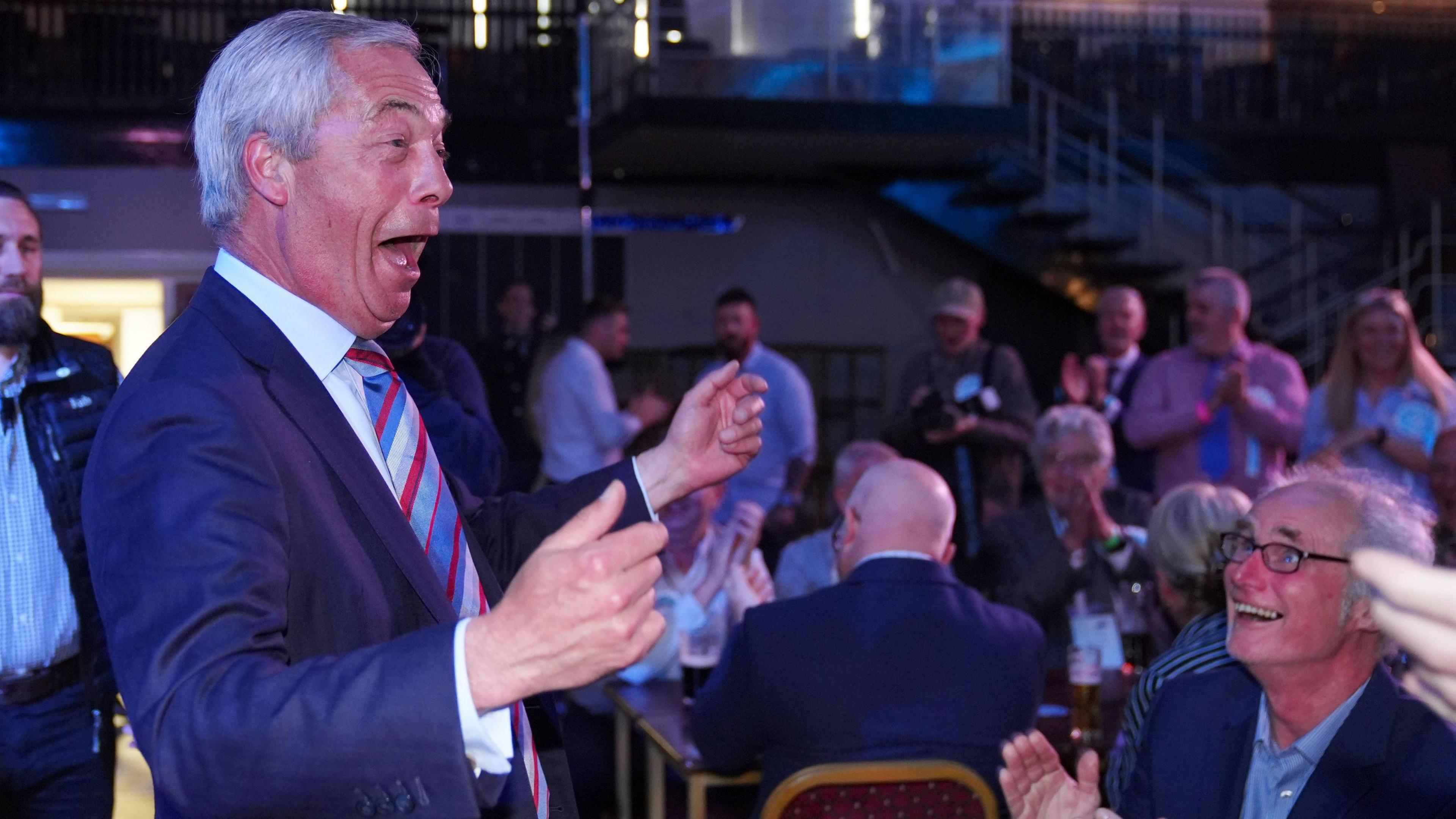 Nigel Farage celebrates by raising his arms in the air as he looks out to a crowd in a venue in Newton Aycliffe, County Durham. People clap and watch him. He's wearing a blue and red striped tie.
