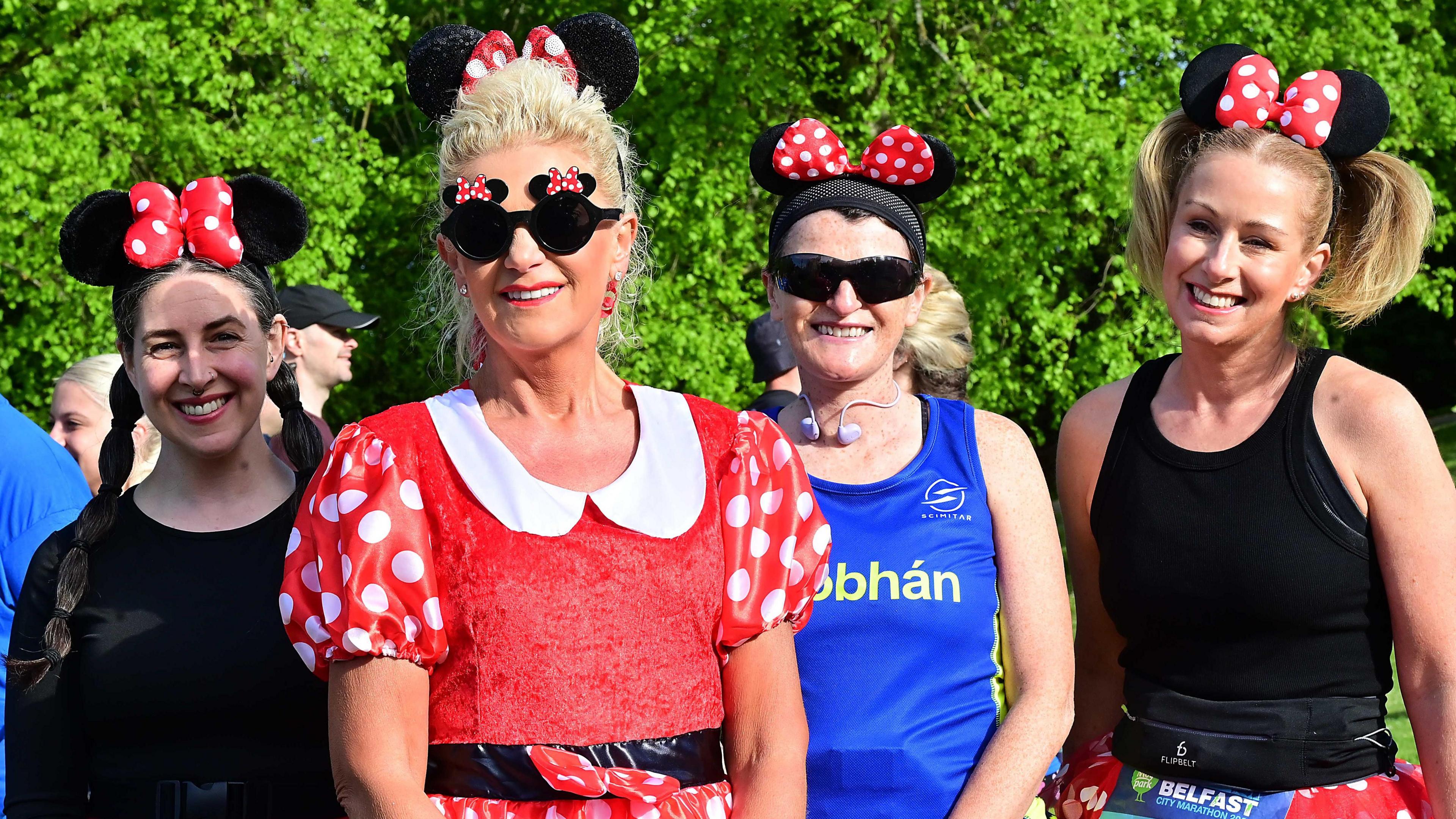 Four women standing next to each other. From left to right the first has plaited long brown hair, black top and is wearing Minnie Mouse ears. Next to her is Elaine who has blonde hair tied back wearing a red Minnie Mouse costume. Next to her is a lady with black clothing and short blonde hair wearing Minnie Mouse ears, and beside her is a lady with a blue top, red shorts black sunglasses and Minnie Mouse ears with her dark hair tied up.