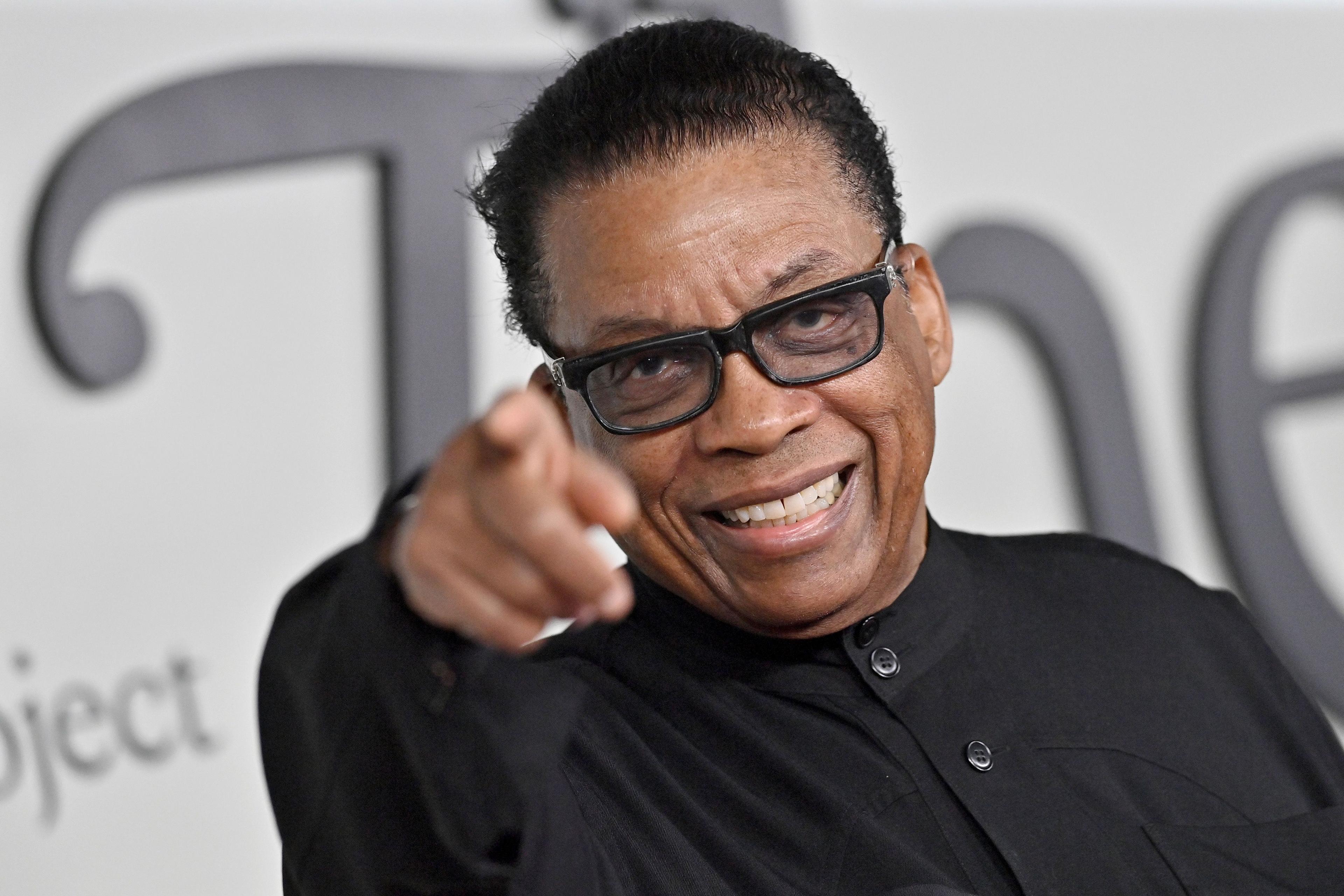 Herbie Hancock pointing straight at the camera. He is wearing black glasses and a collarless black shirt.