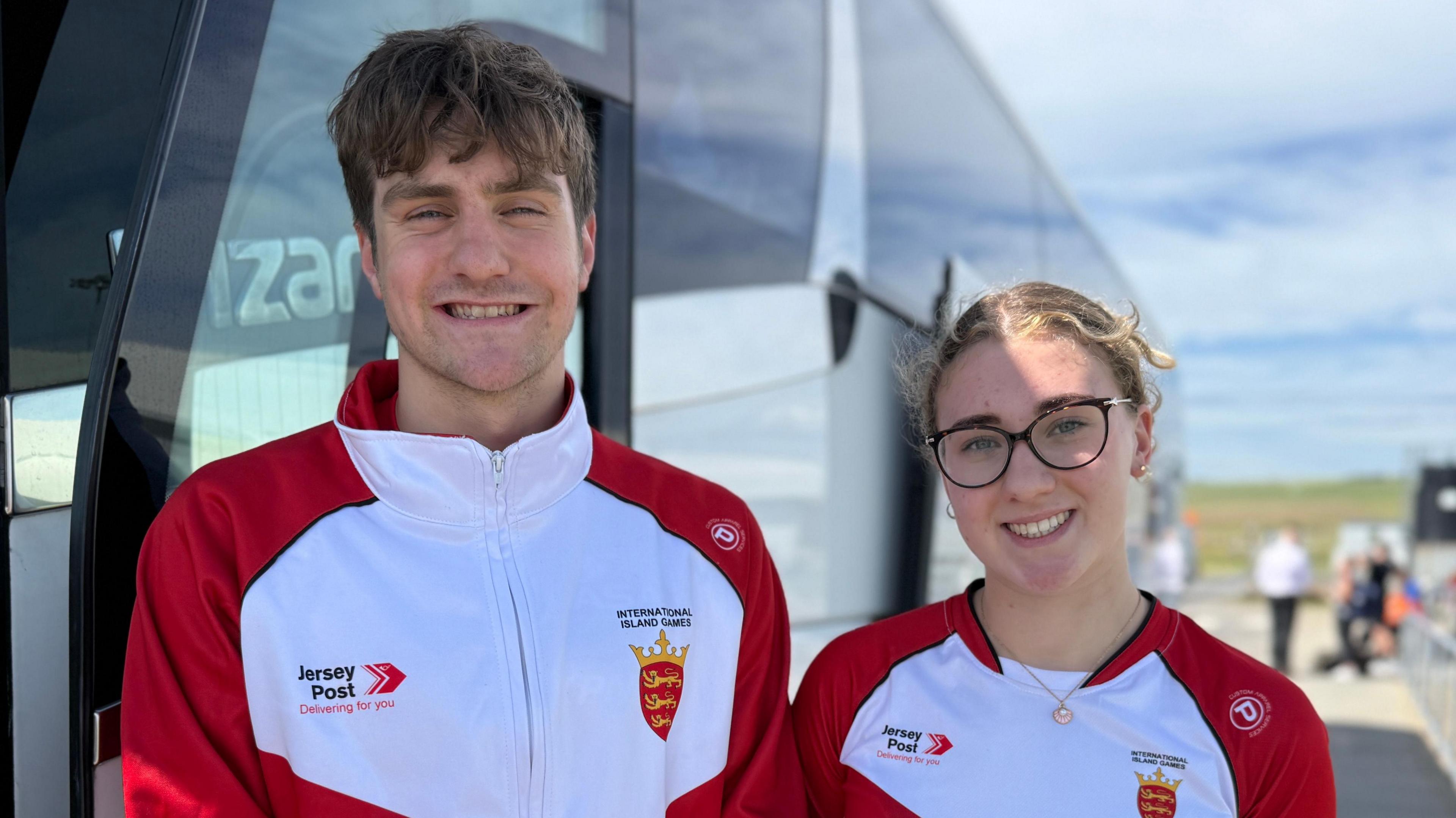 Isaac Dodds and Megan Hansford are standing in front of a coach smiling into the camera, they are both wearing red and white Jersey sports kit.
