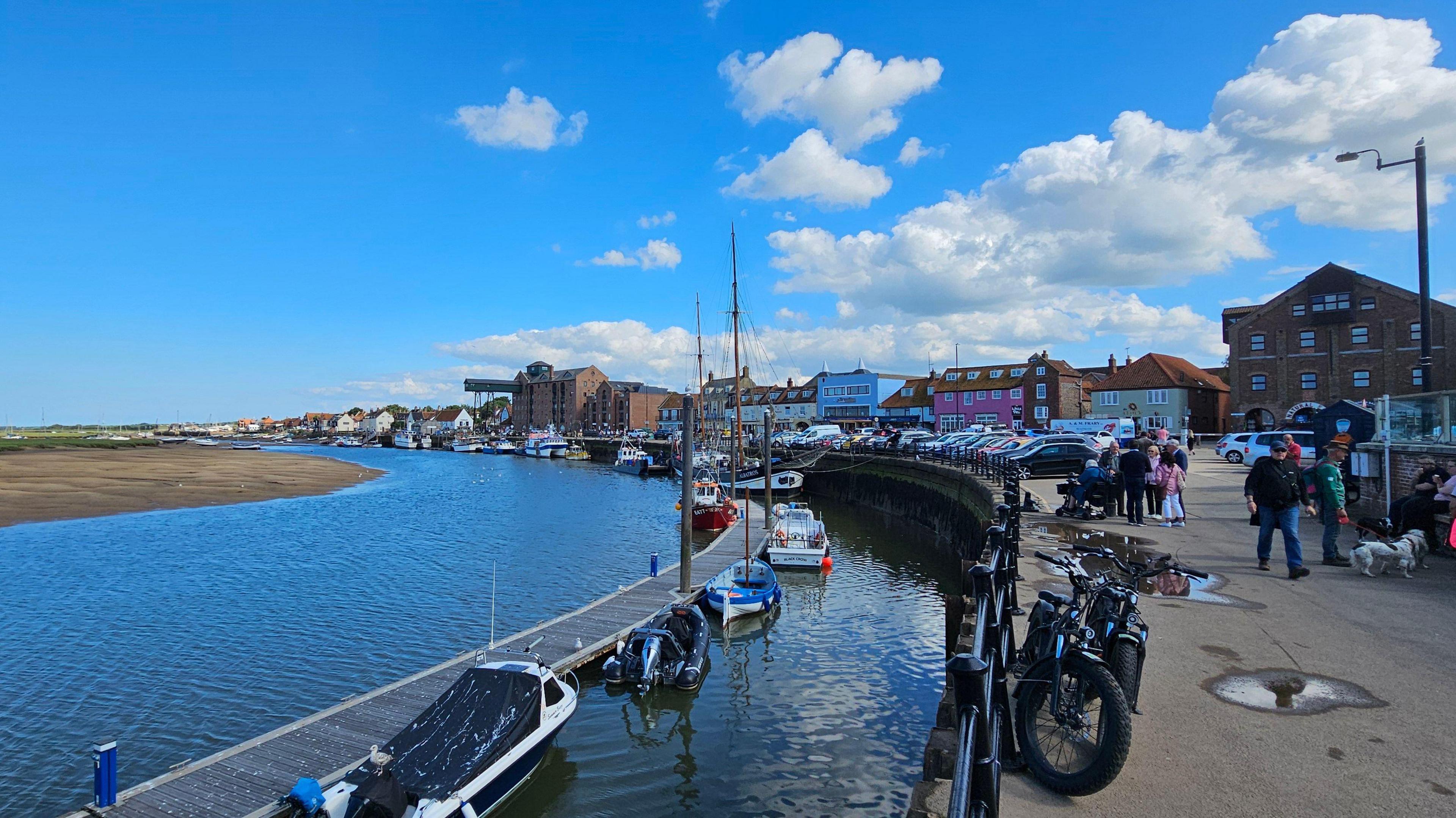 The quayside at Wells-next-the-Sea, with boats moored along a jetty. On the quay, there are mountain bikes parked and groups of people huddling. There are some colourful Victorian houses and buildings in the background. The sky is blue, with only a few white clouds.