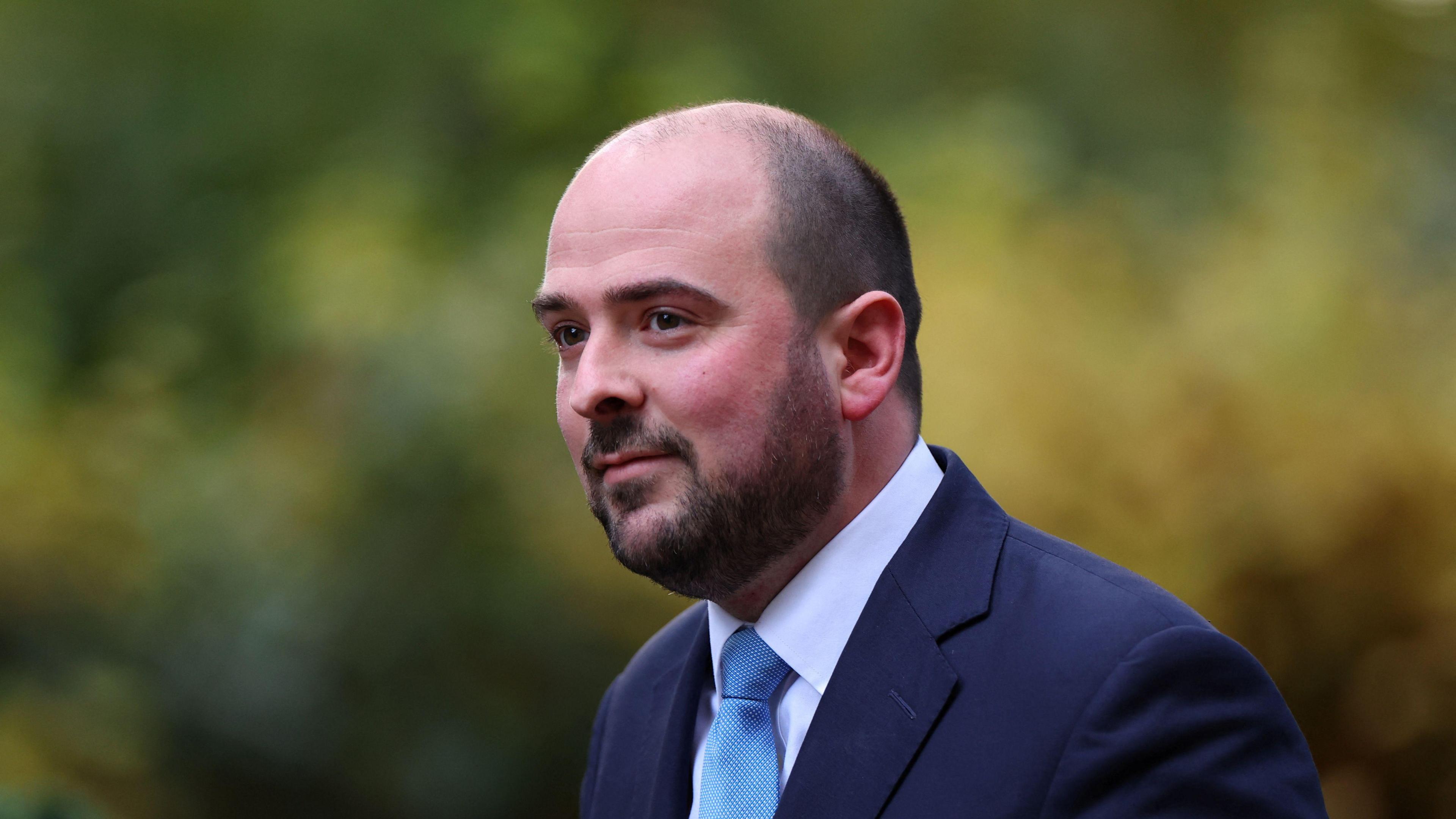 A head-and-shoulders picture of MP Richard Holden, a bald man with a dark brown beard. He is wearing a navy suit with a white shirt and blue tie.