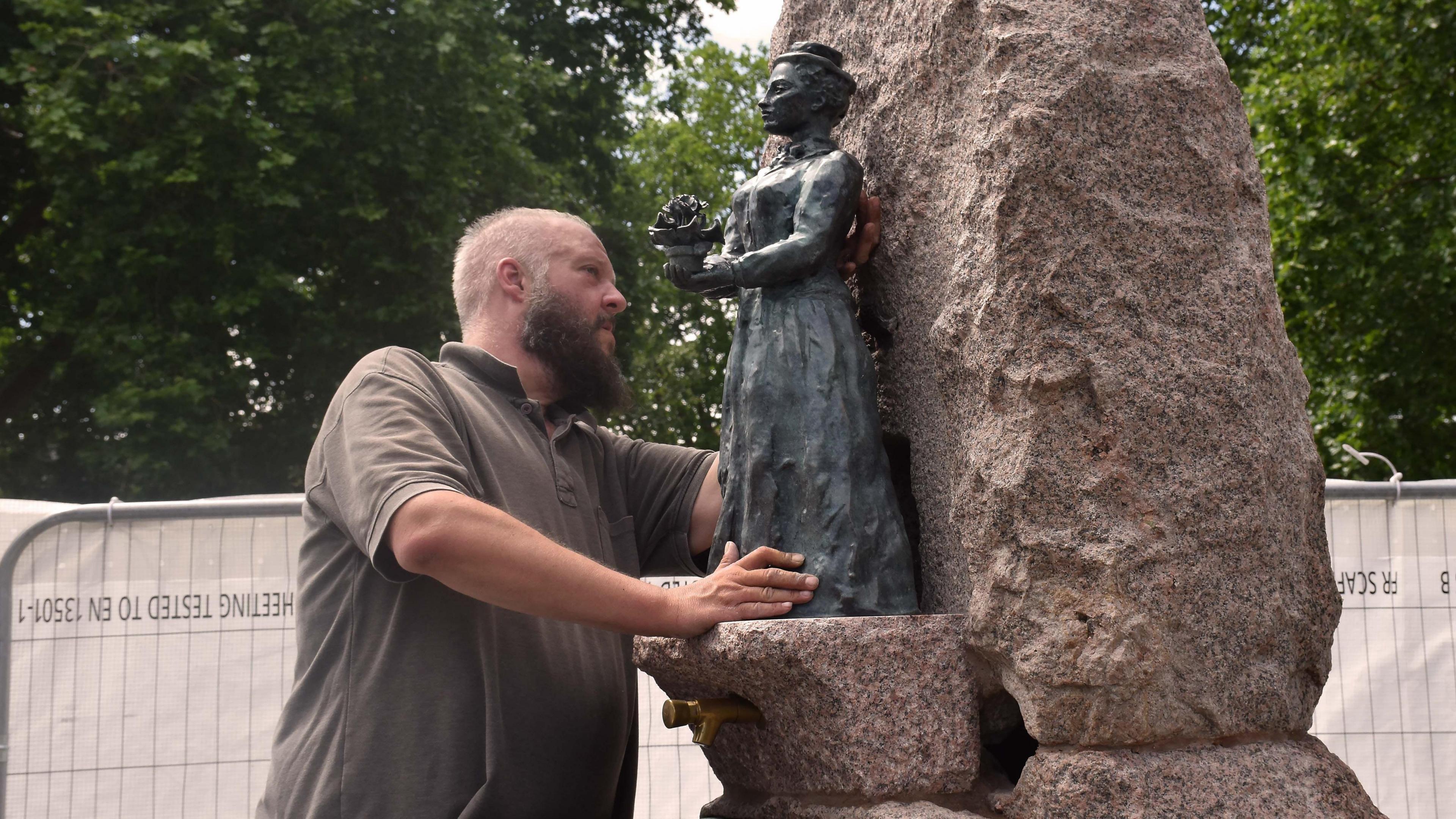 A man in a brown T-shirt installs a bronze statue on a large rock-style water fountain. There are tall trees in the background.