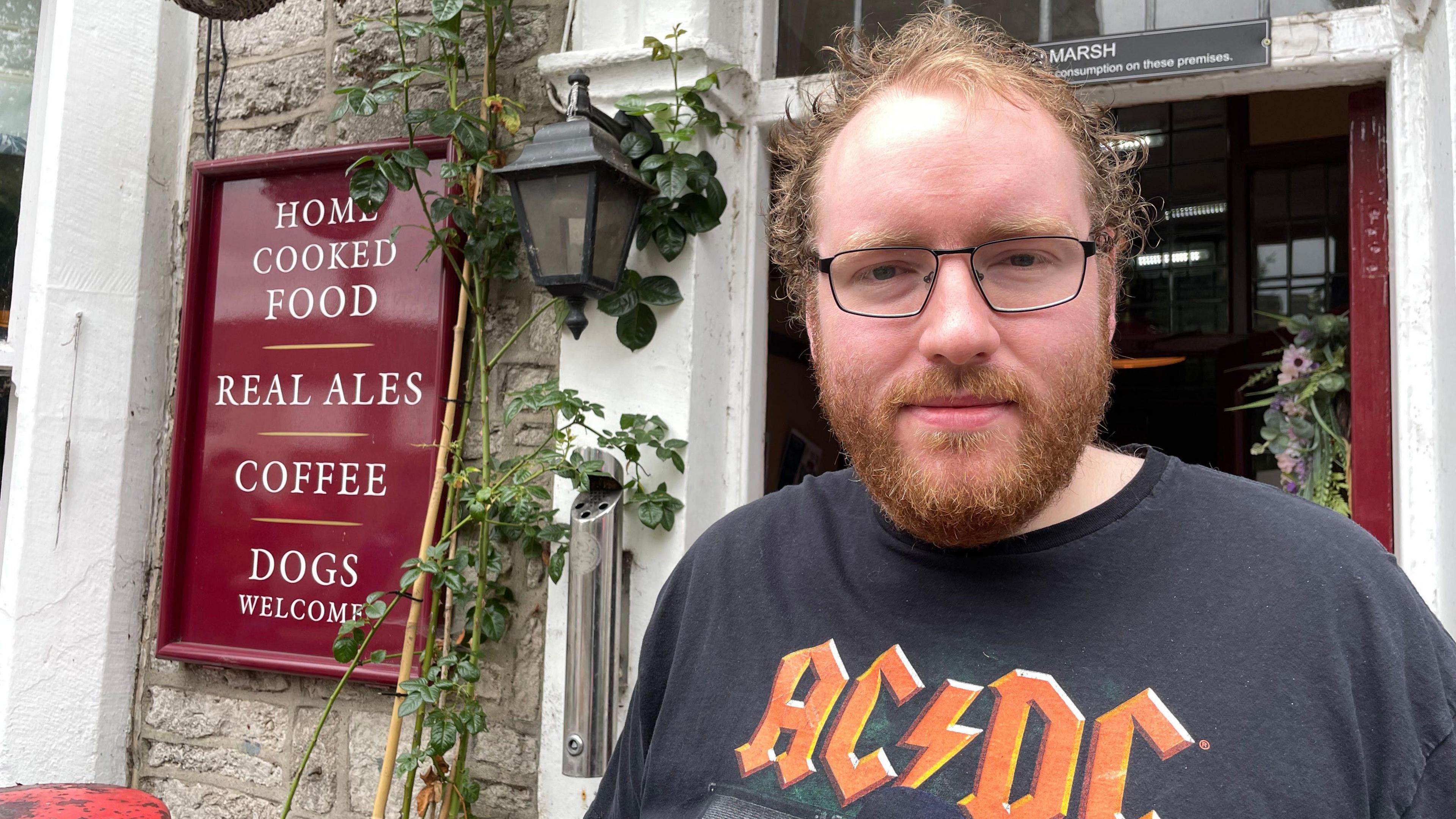 Man with a beard and glasses wearing an AC/DC T-shirt in front of a pub sign