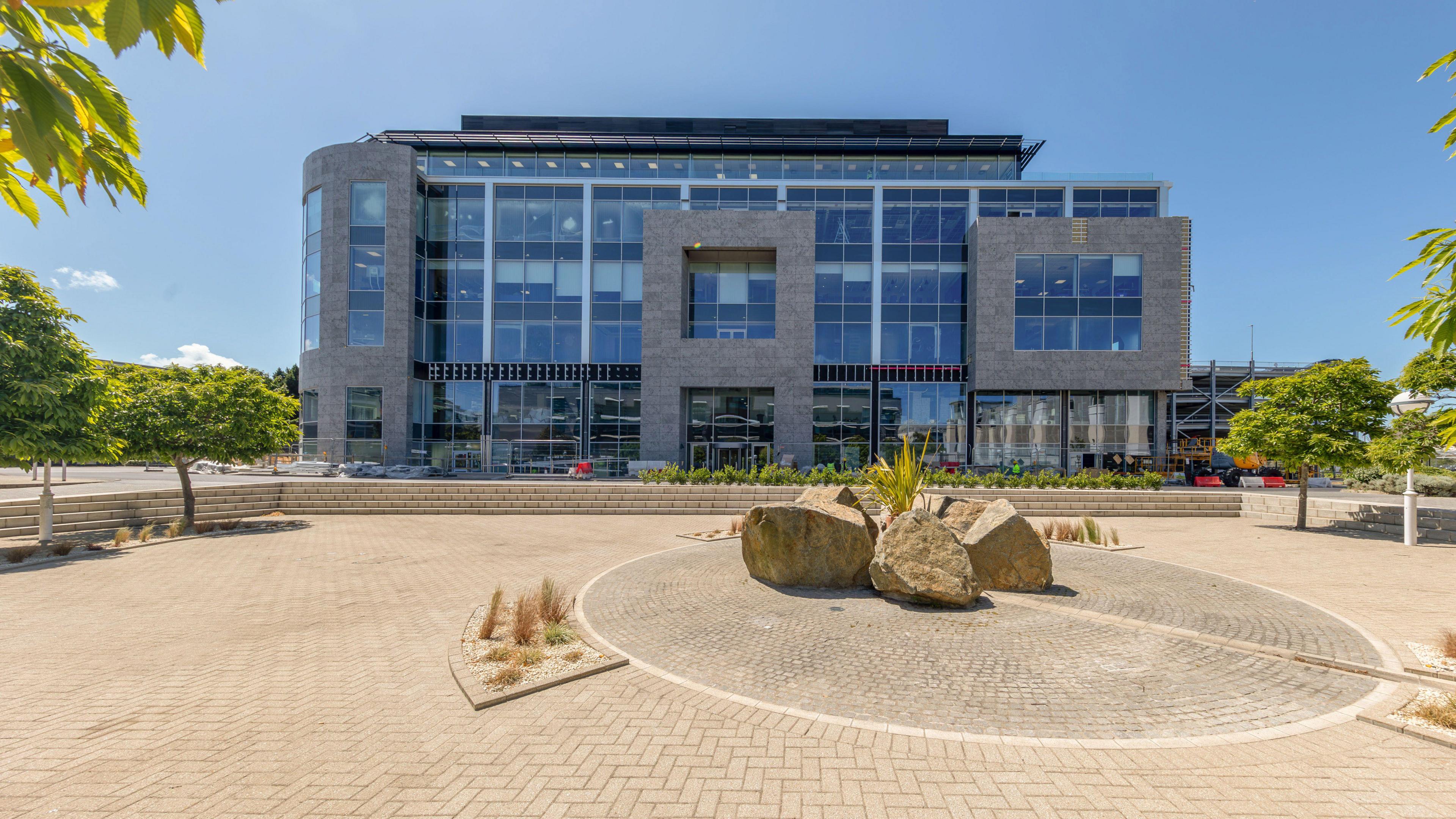 A large grey building with a wall of blue windows. There is a paved area at the front with large beige rocks