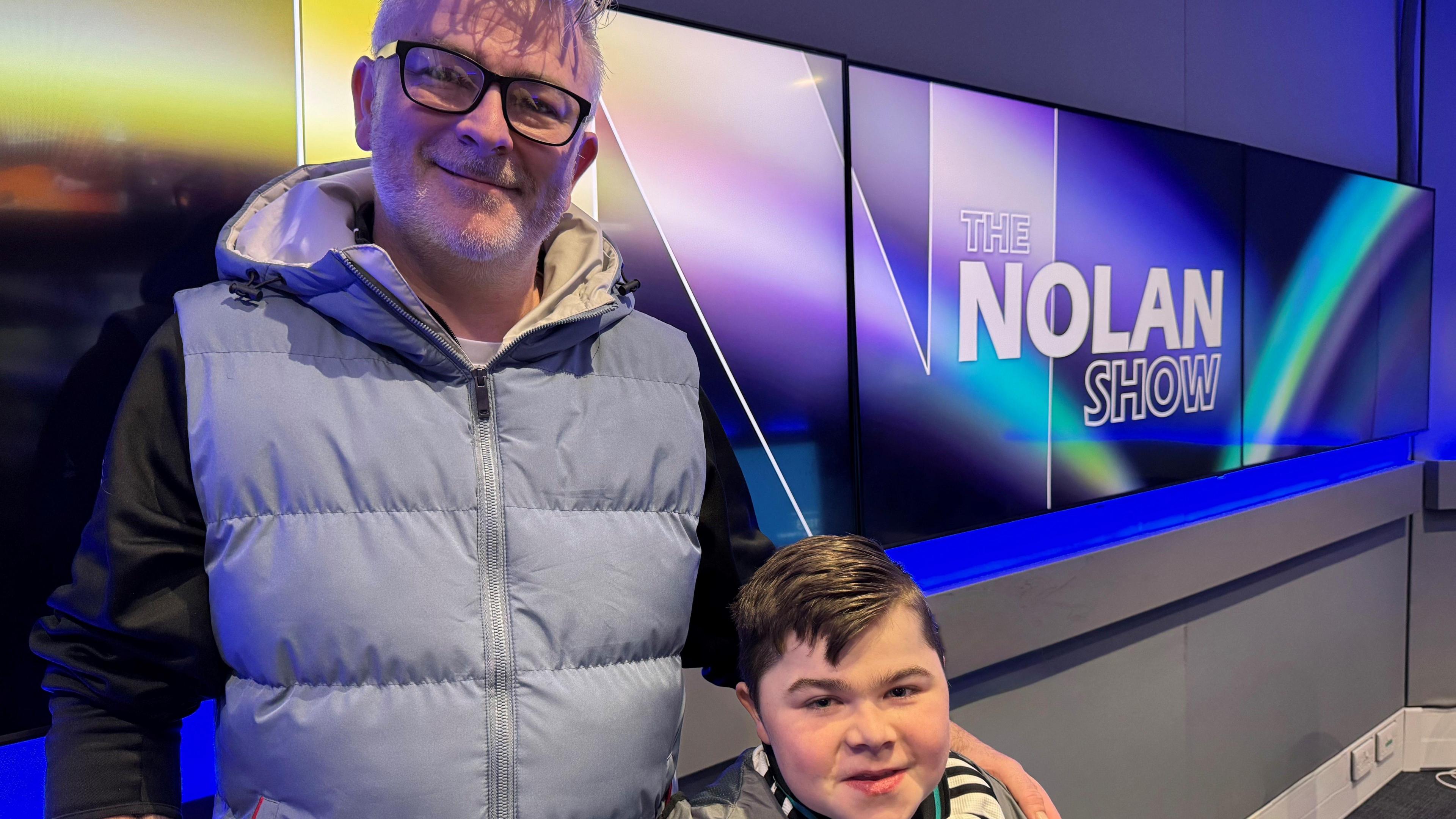 Jamie with his arm around Alfie. Jamie is wearing glasses and a light blue puffer gilet with black top underneath. Alfie has dark hair and wearing a football jersey. A screen with The Nolan Show branding can be seen to the right.