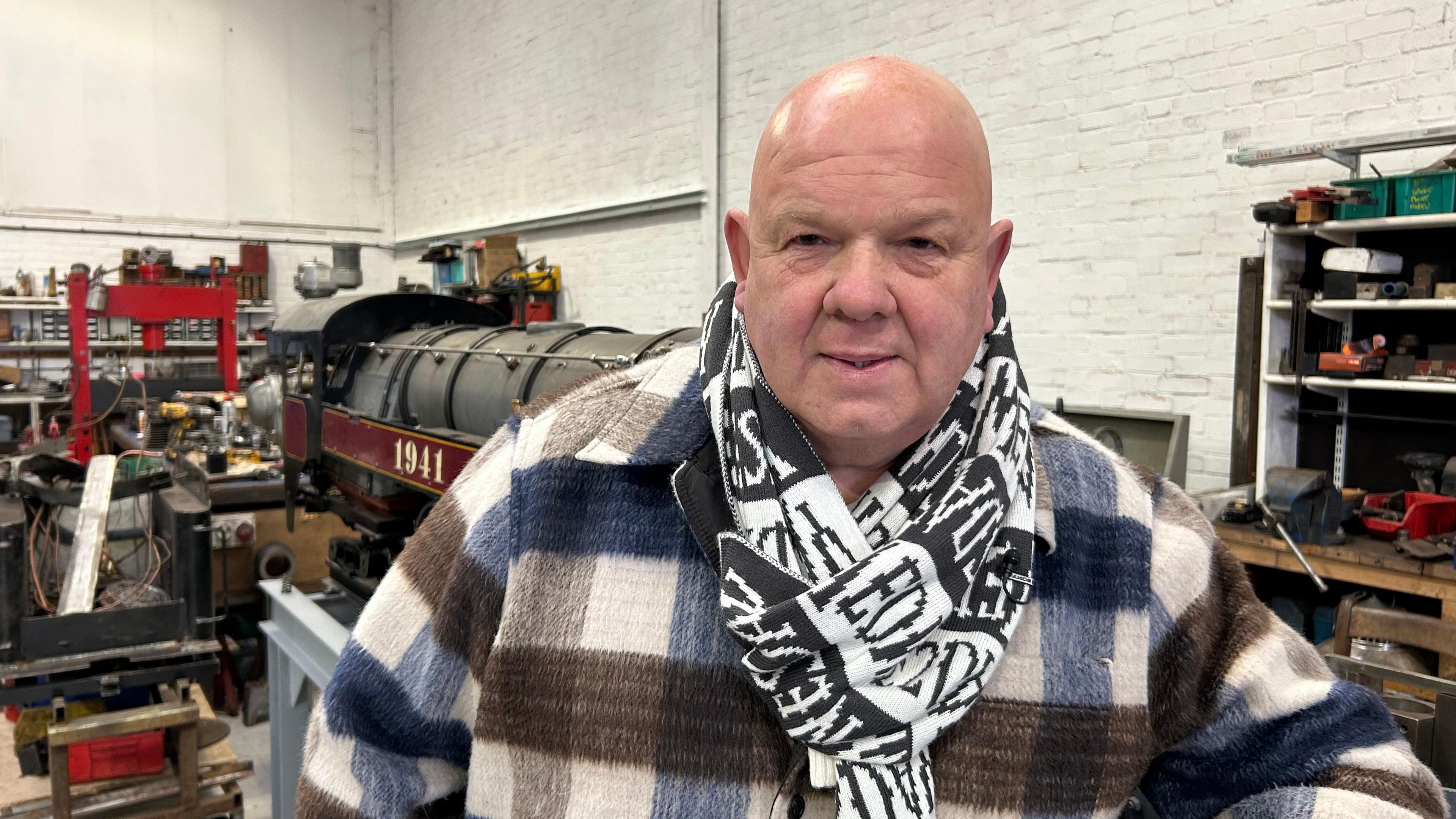 A man wearing a checked flannel shirt and a black and white scarf, standing in a workshop. A miniature train engine can be seen in the background alongside walls and work benches filled with tools and train parts.