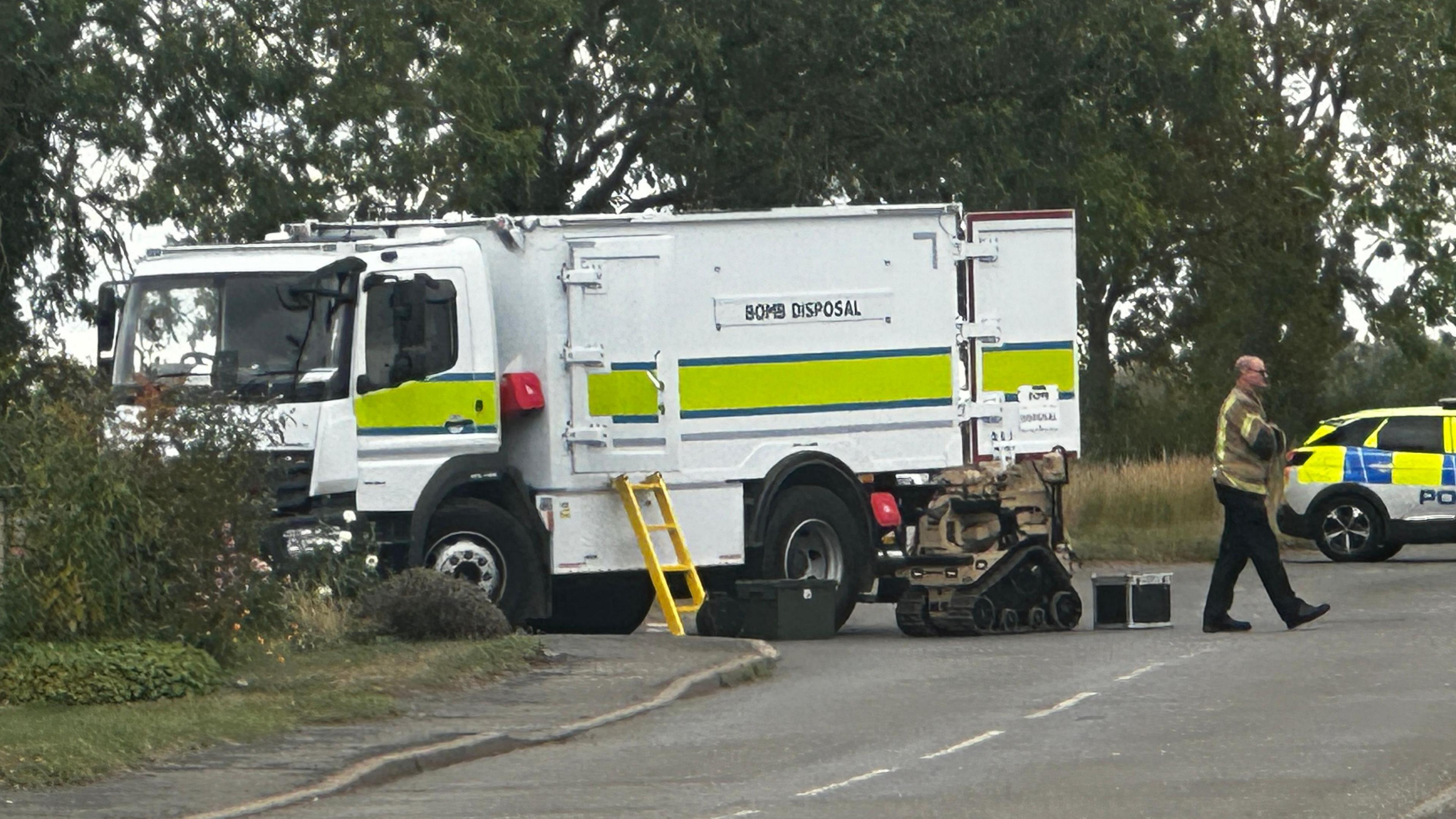 White truck with yellow and blue stripe. Bomb disposal written on it. There is a yellow ladder, a bomb disposal robot, a police car and a man in police bomb disposal officer. There are trees, bushes, a road and a path.