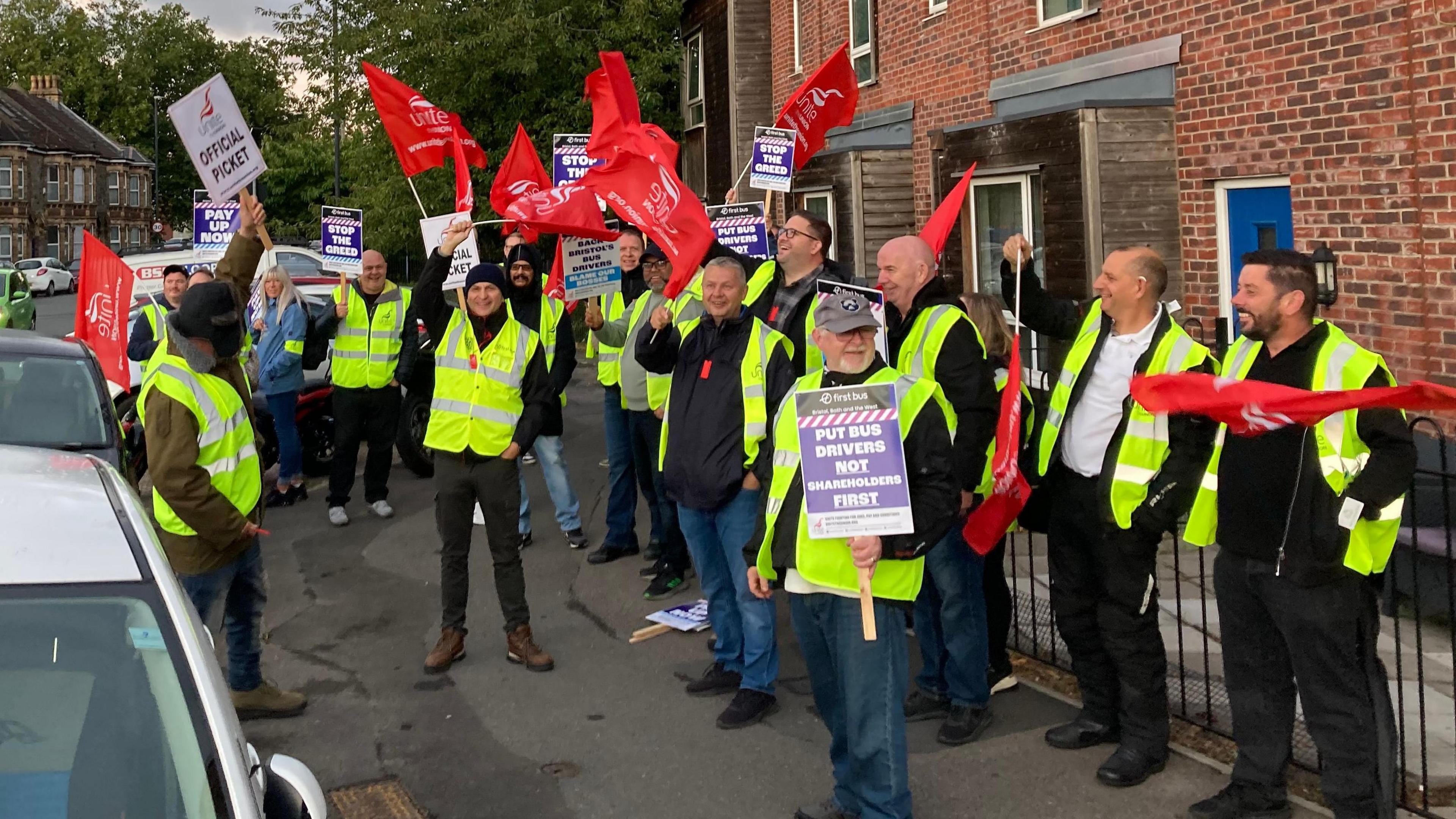 A large group of First Bus Drivers, wearing yellow hi-vis jackets and holding up red Unite union flags and placard signs demanding fair pay. They are standing on a wide pavement outside a red brick building, beside a row of parked cars. 