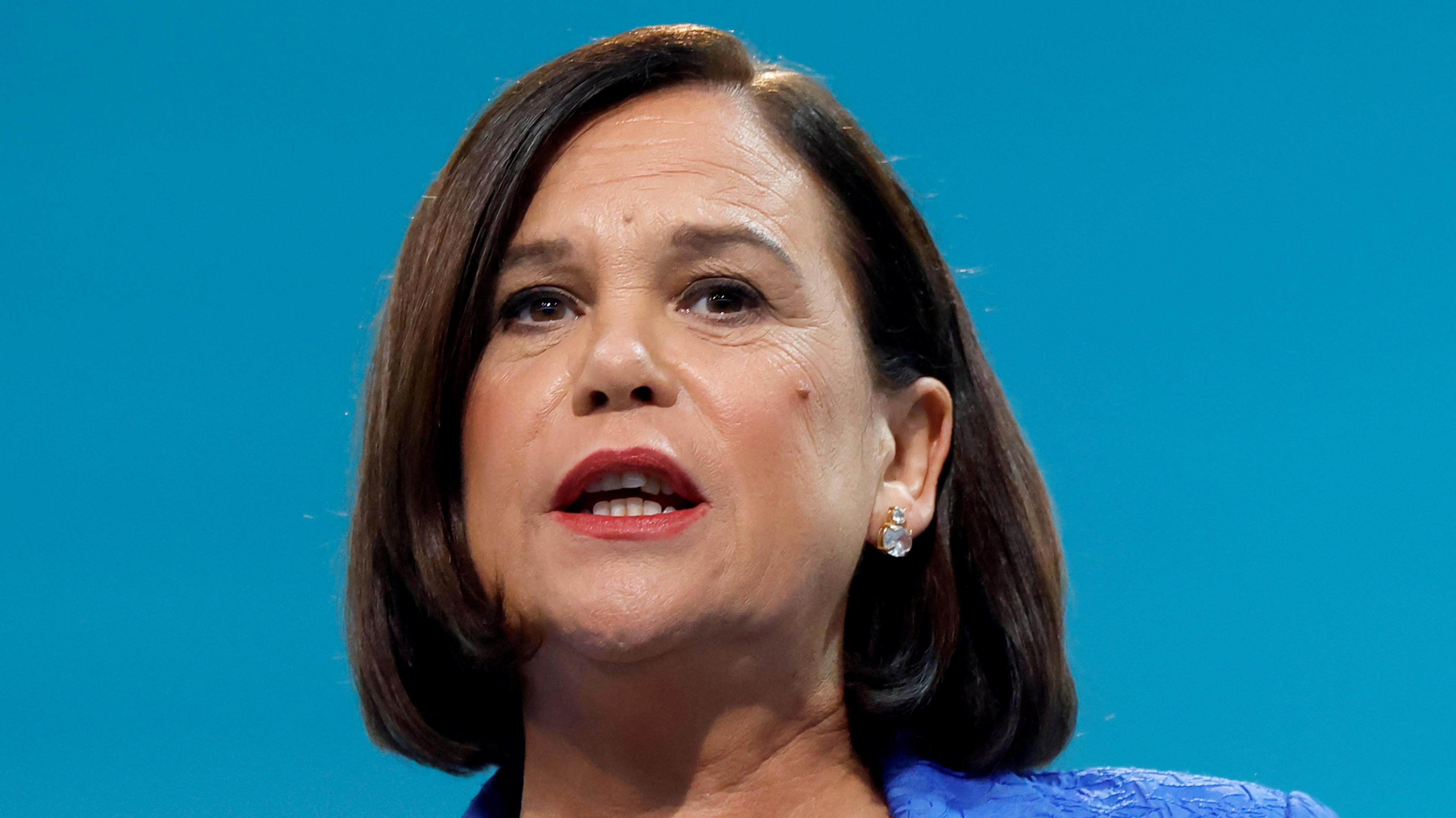 Mary Lou McDonald, who has a brown bob-type hairstyle and wears earrings and a blue jacket as she speaks on a stage.