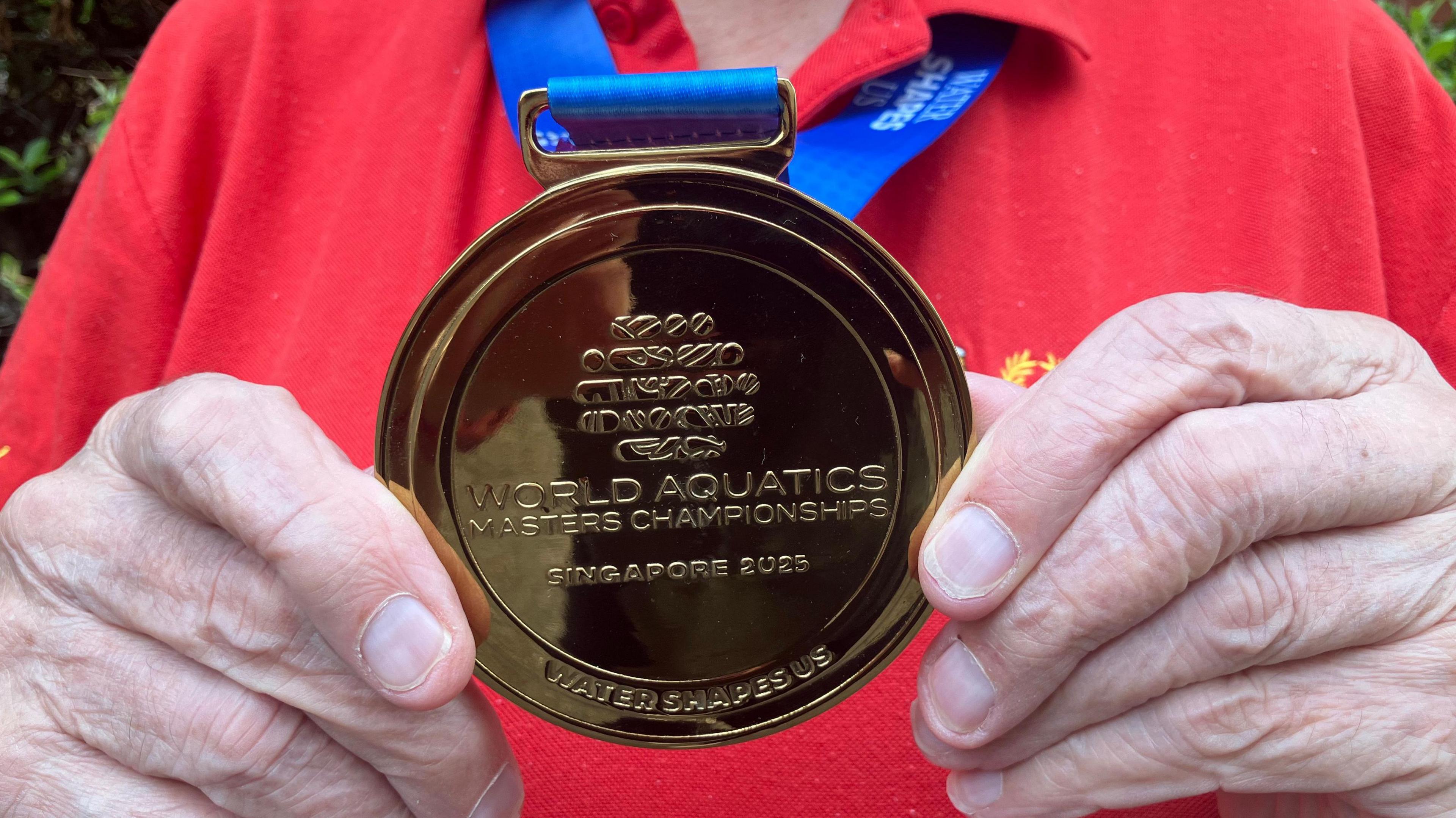 A close up view of Mr Read's gold medal as he holds it. Engraved on the medal are the world World Aquatics Masters Championship Singapore 2025.