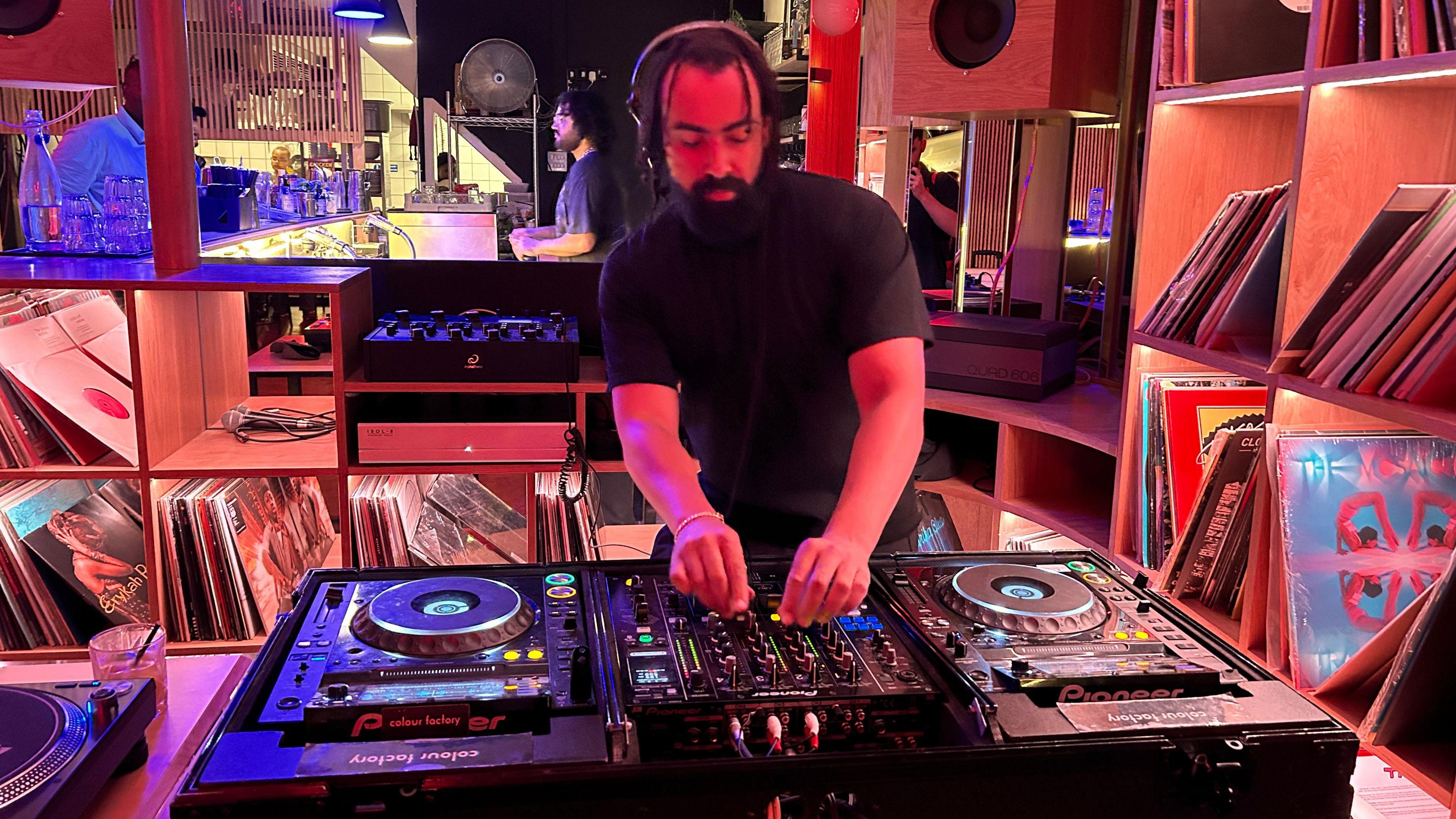 A man wearing over-ear headphones tinkers with the controls of a mixing deck. He's surrounded by shelves of vinyl records. He looks as if he's concentrating very hard.