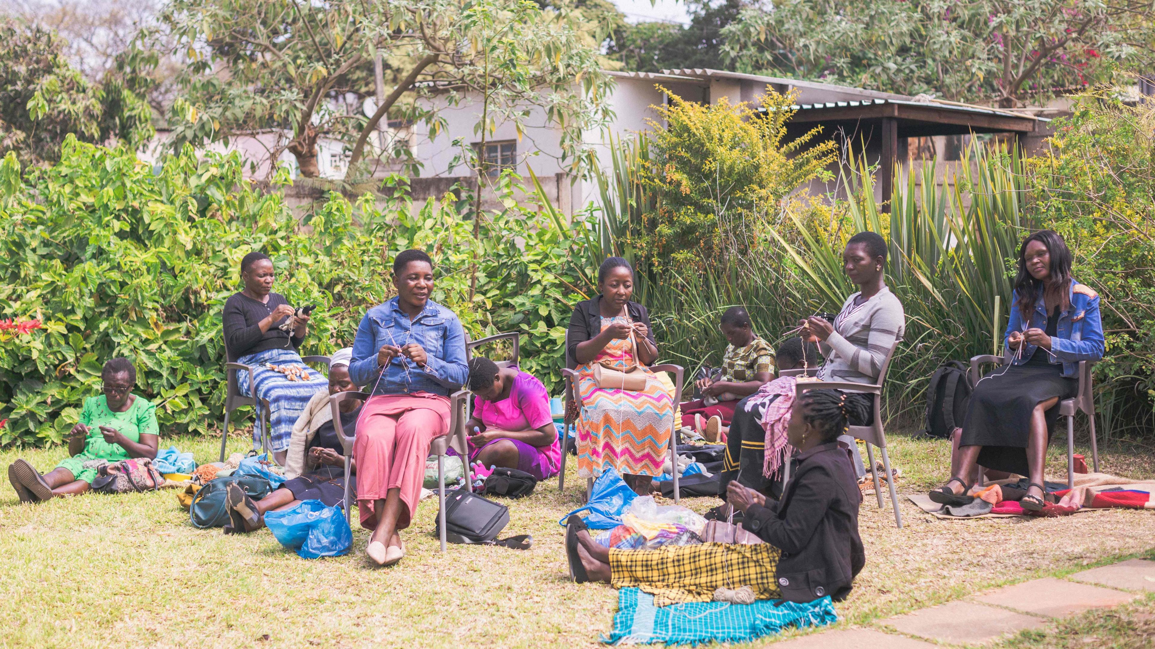 A group of 10 women knitting in a garden. Five are sitting on chairs; the rest sit on rugs spread across a lawn. There are shrubs and trees in the background. 