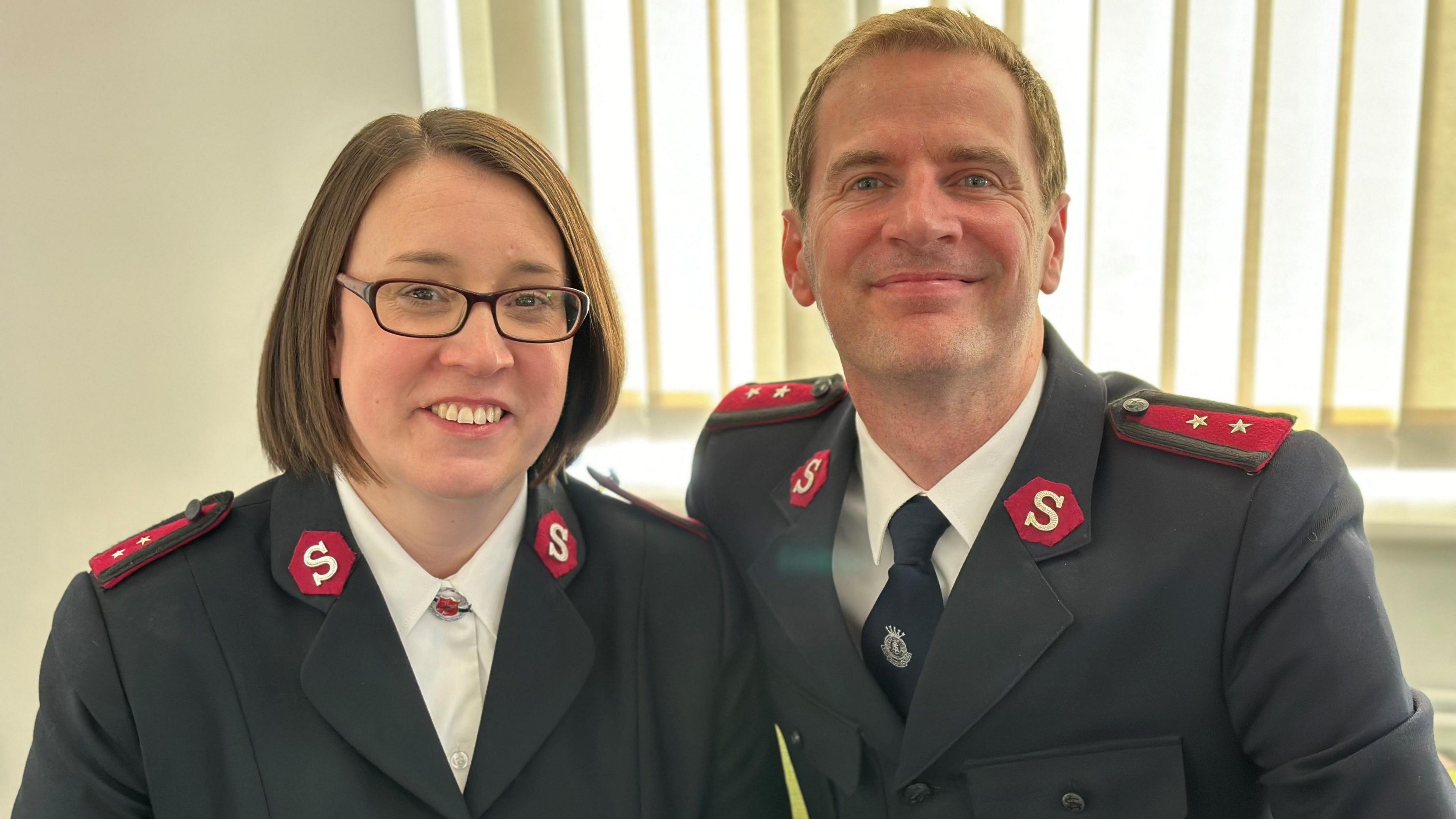 Rachael and Dylan Nieuwoudt in their Salvation Army uniforms smiling at the camera. The uniforms are black formal jackets with red epaulettes featuring stars in the shoulders and red hexagons featuring the letter S on the lapels. Both are wearing white shirts with the collars buttoned up. He wears a dark tie with a white emblem and she wears a broach. 