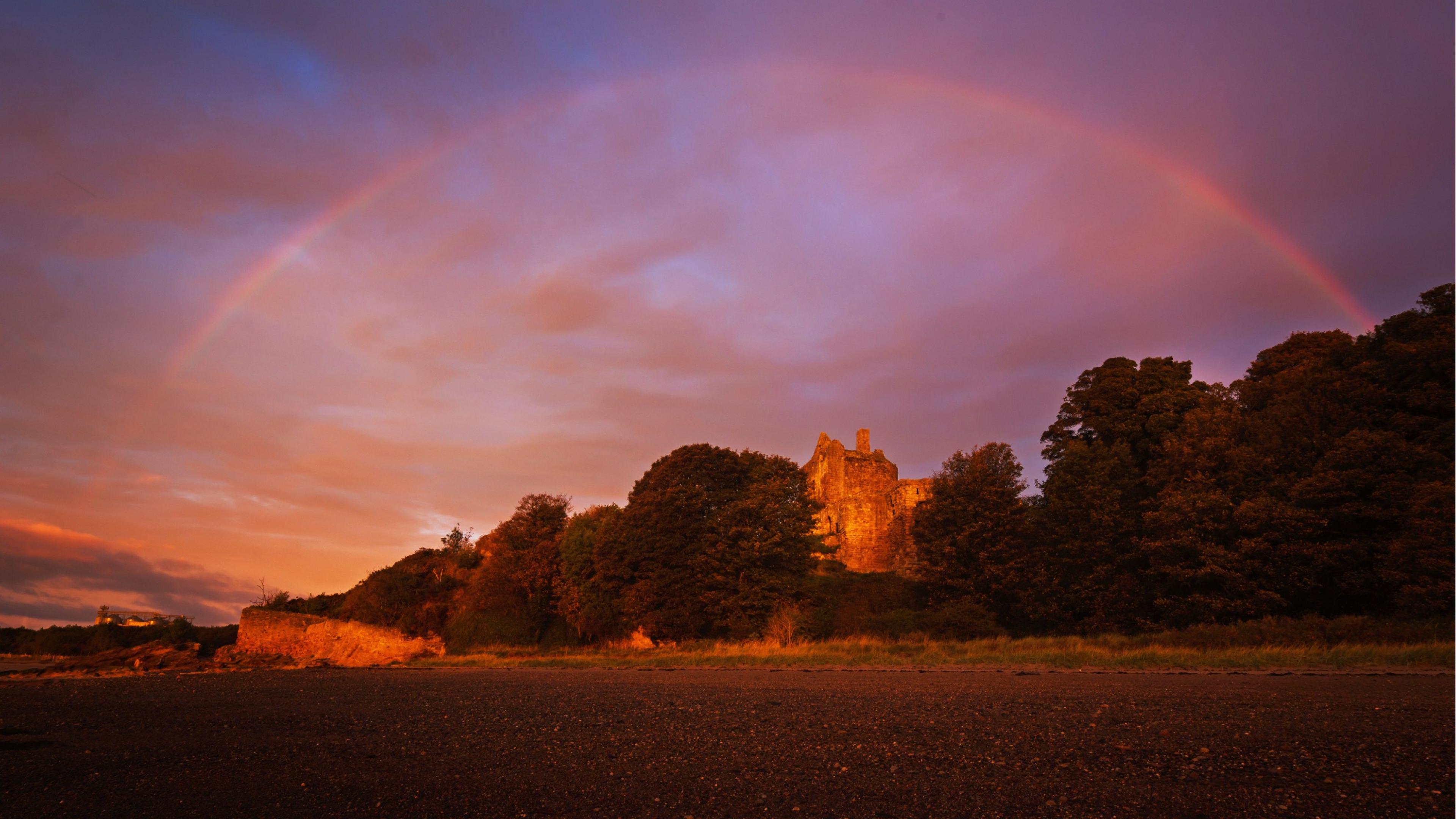 Landscape of a wooded area with the rooftop of a castle visible, below a vibrant purple, pink and orange sky with a rainbow above the castle and trees.