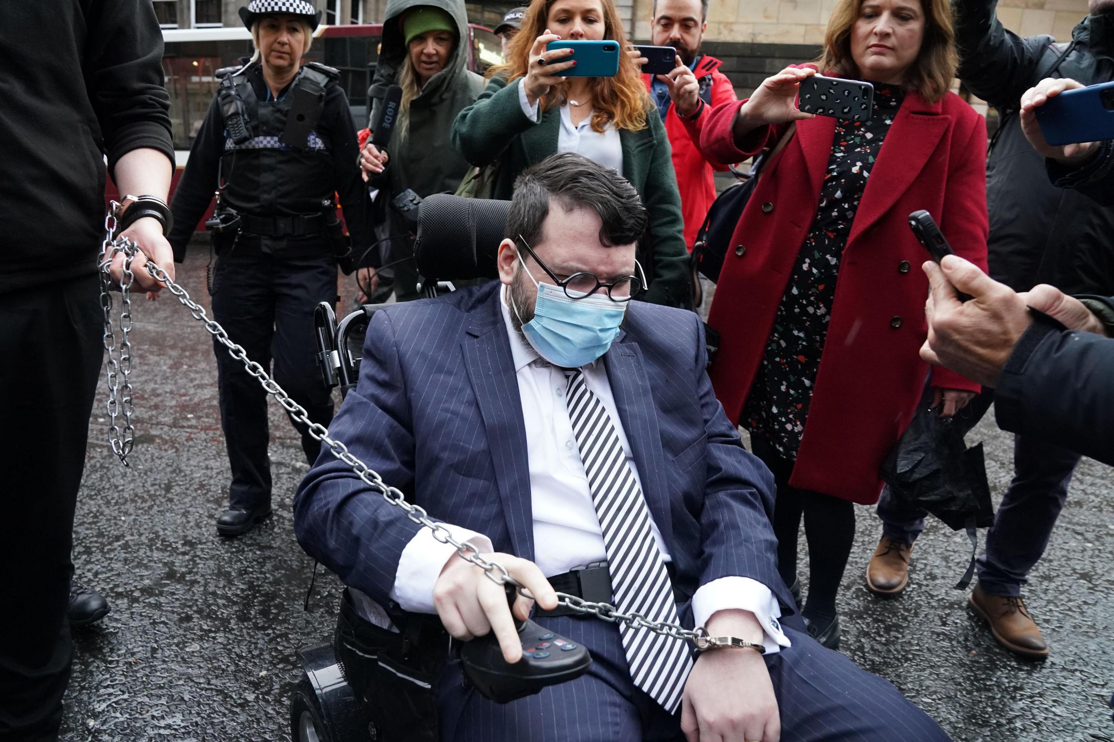 Nicholas Rossi outside court in Edinburgh. He is in a wheelchair and is wearing a blue suit, white shirt, striped tie, glasses and an oxygen mask. People behind him are filming him on their phones and a long chain being held by a police officer links to his handcuffs.