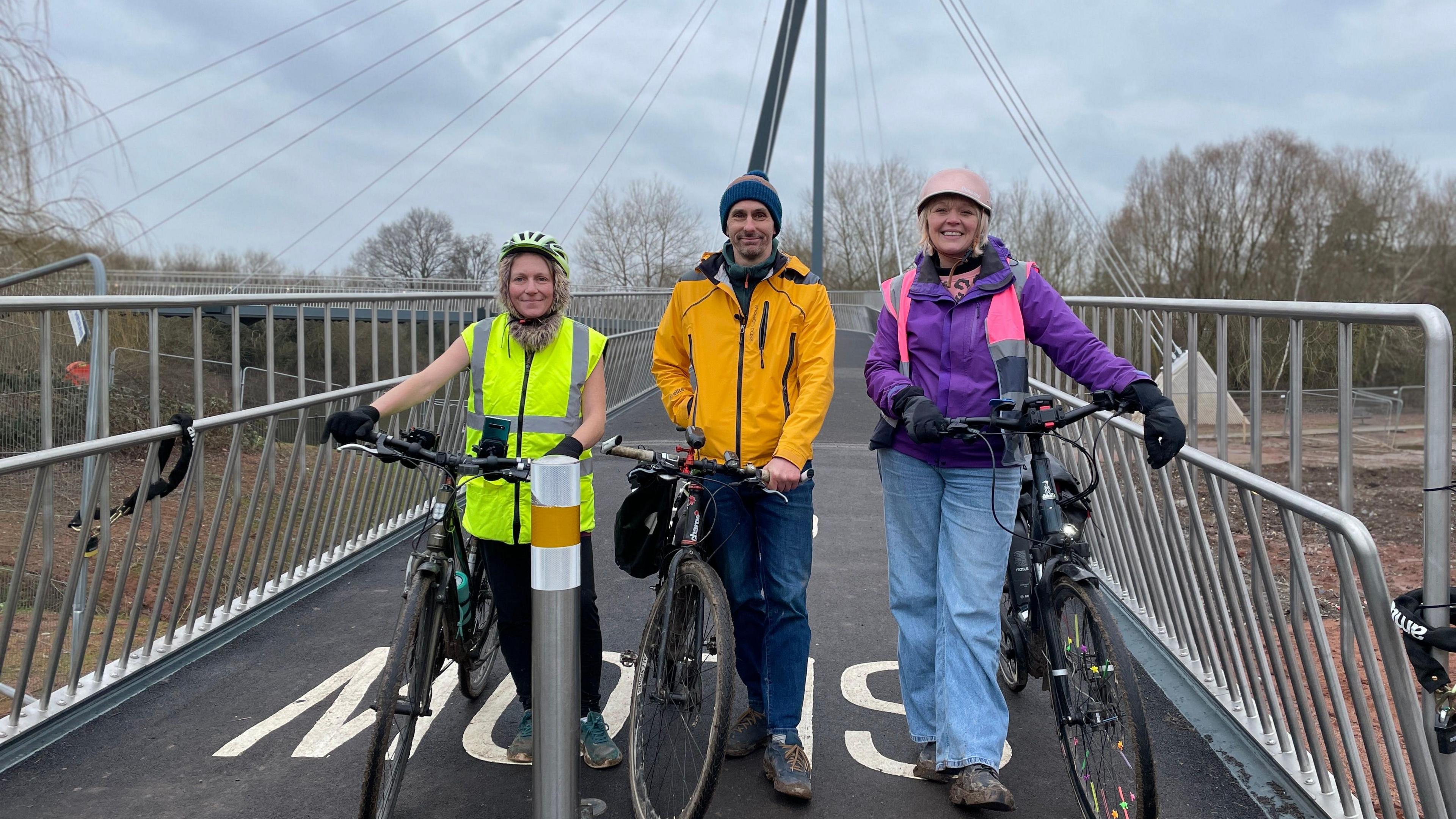 Three people - a woman wearing a hi-vis and black trousers and yellow cycle helmet, a man wearing an orange jacket, dark blue jeans and a beanie, and a woman wearing a purple coat, pink and grey hi-vis, light blue jeans and pink cycle helmet - stand next to their bikes on the Kepax footbridge, with temporary barriers either side of them.