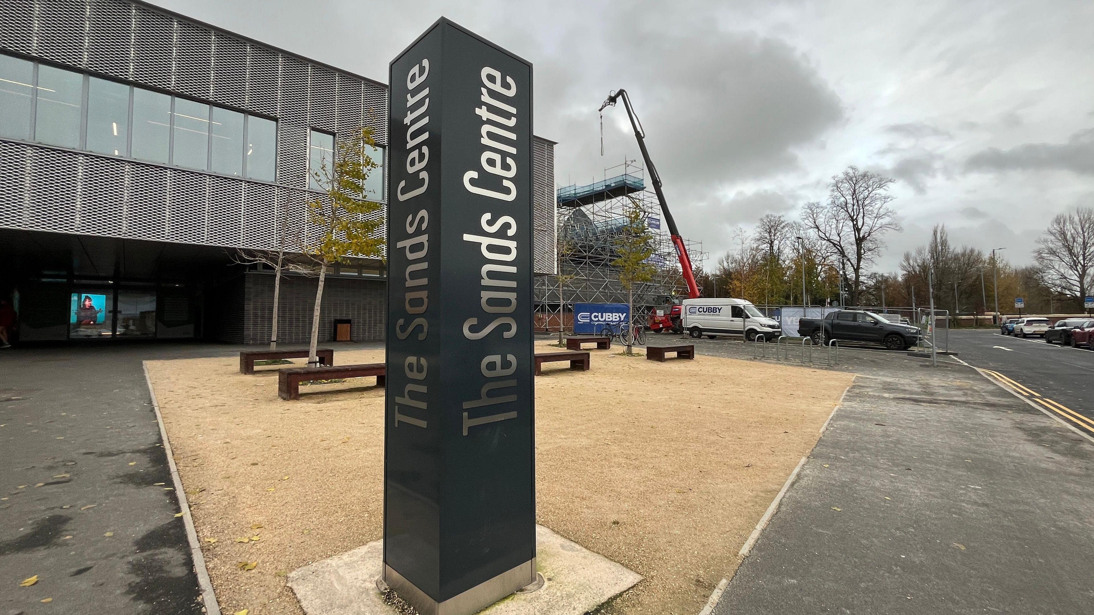 A general view of the entrance to The Sands Centre in Carlisle. A black pillar has the name of the venue written on each side in silver. The entrance to the leisure centre is visible on the left, while on the right heavy plant machinery is working on an area of the building surrounded by scaffolding.
