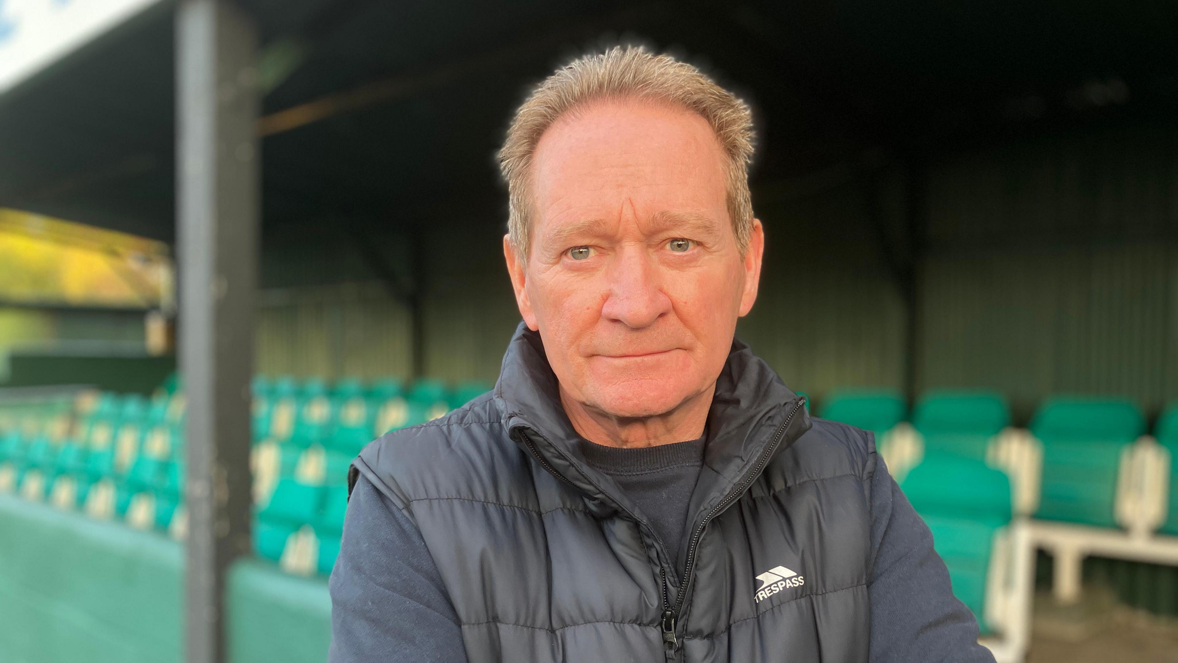 A middle-aged man stares at the camera whilst wearing a dark coloured gilet and pullover. He stands in front of a football club stand made up of green tipper seats.