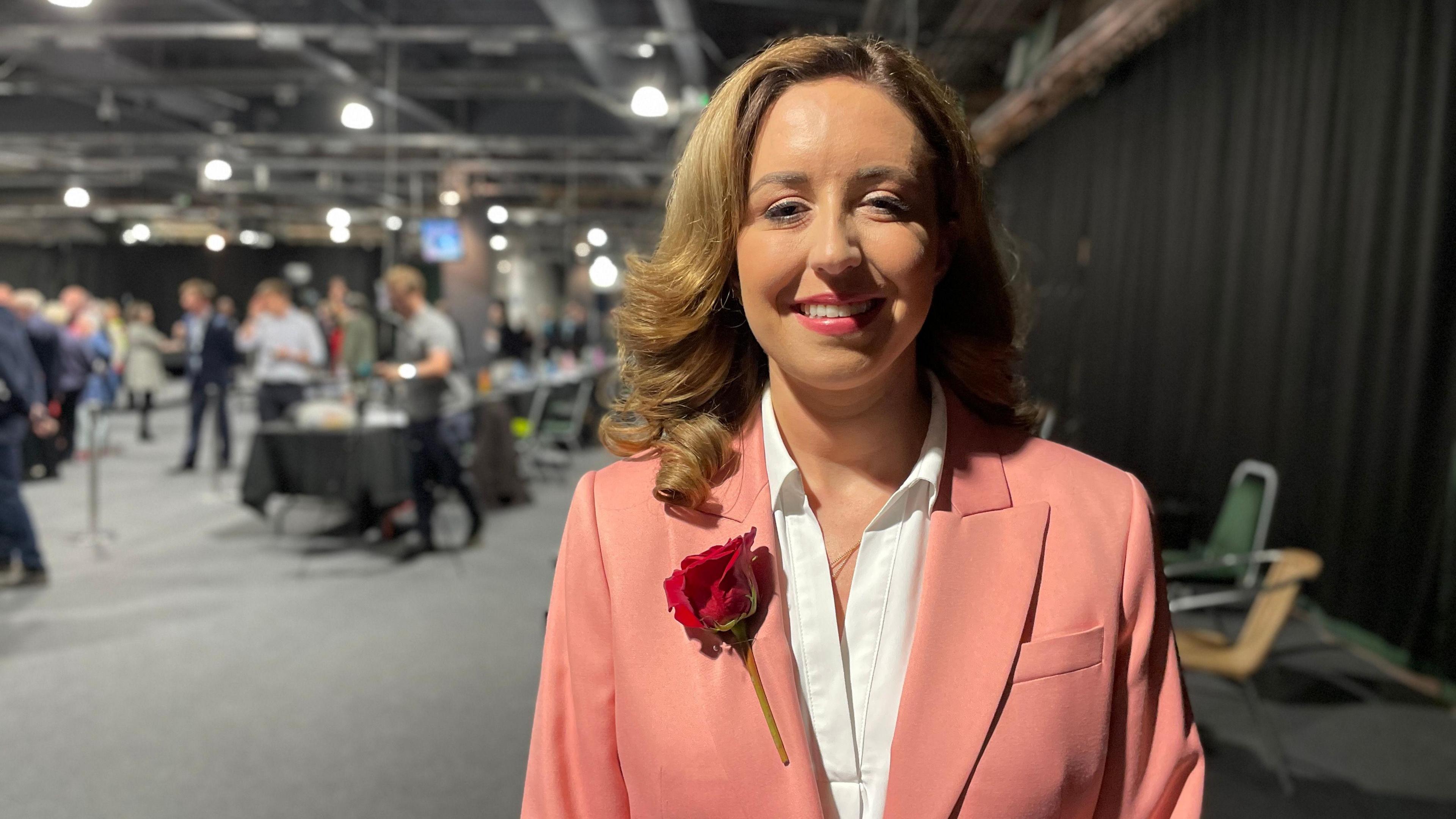 A woman with shoulder length light brown hair is wearing a white shirt and a peach coloured blazer with a red rose pinned to the lapel. She is stood at an election count and is smiling