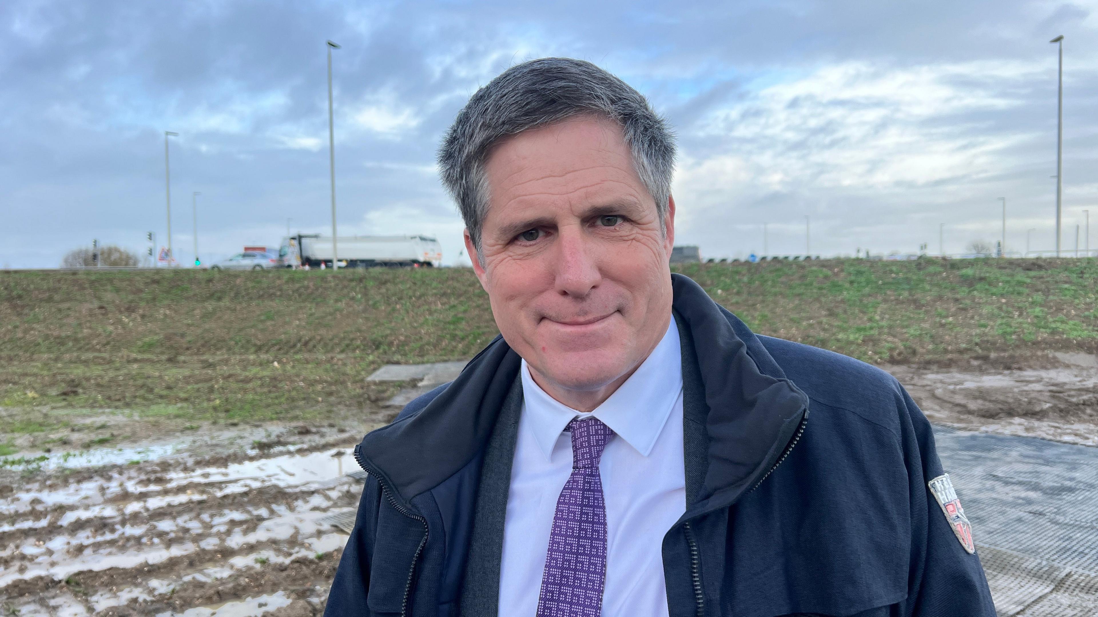 Anthony Browne is standing next to a muddy field. He is looking directly into the camera. He is wearing a light blue shirt, purple tie and navy waterproof jacket.