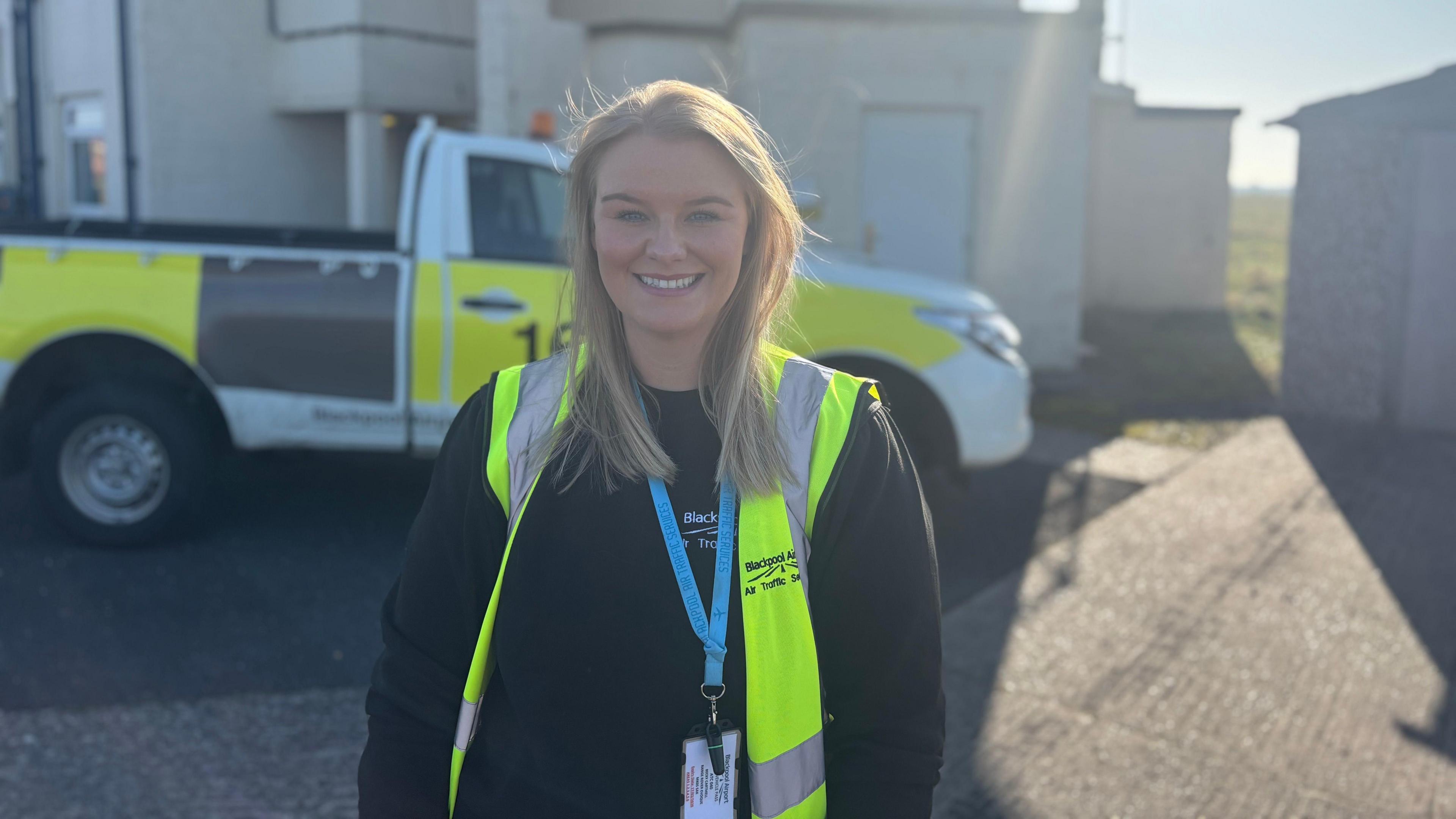 Becky stands in front of the air traffic control building at Blackpool Airport. She has blonde hair and smiles at the camera. She wears a black jumper and a yellow high-vis vest.