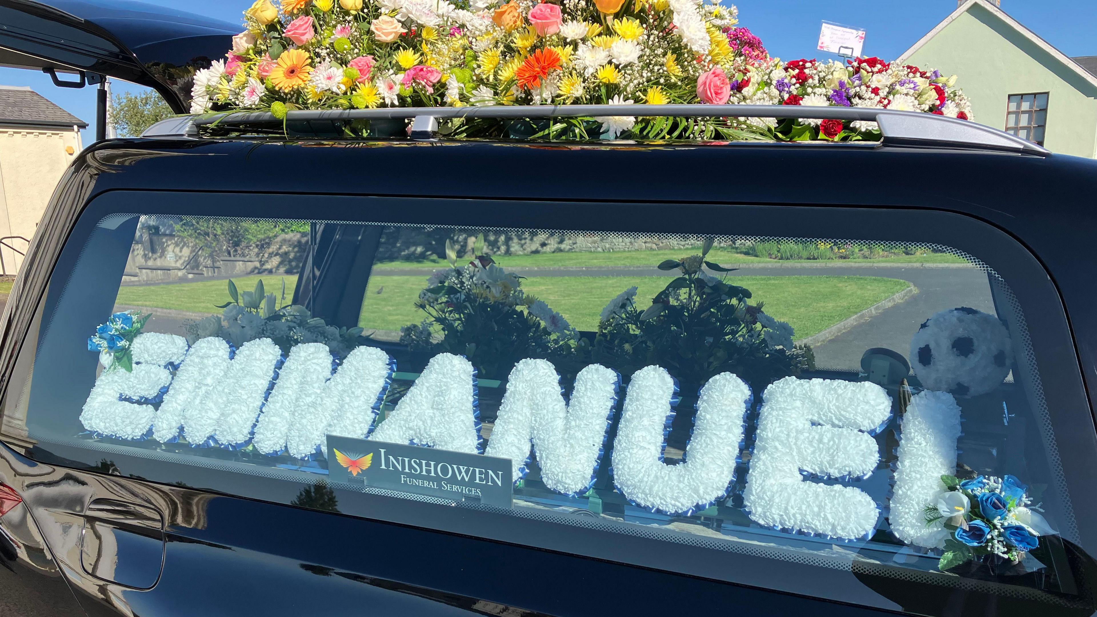 A black hearse with Emmanuel's name printed in white in the window. There are colourful summer flowers on the roof of the hearse.