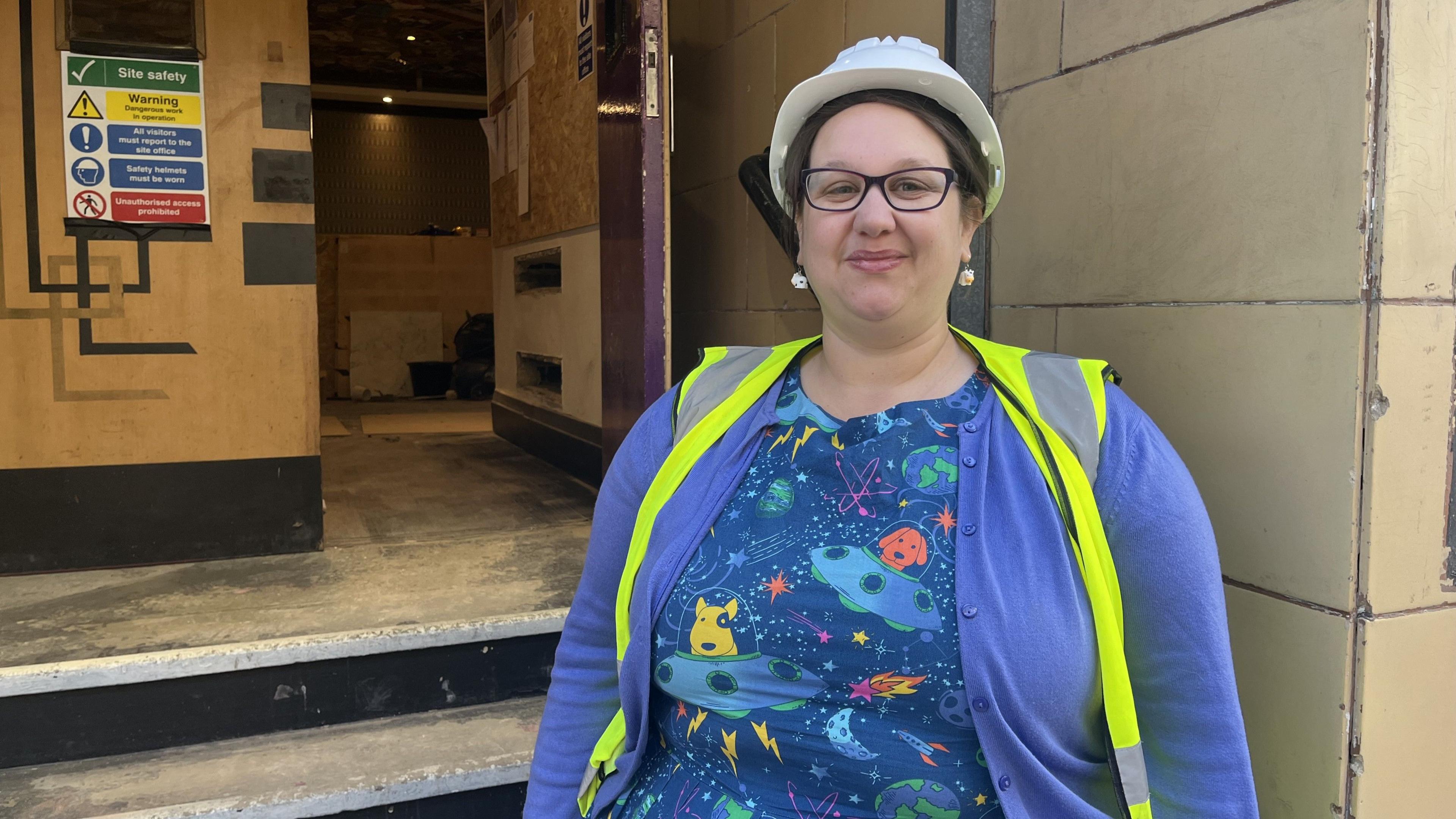 Megan Wilson wearing a blue cardigan, navy dress, high-vis jacket and white hard hat stands at the bottom of the steps leading into the Bradford Playhouse