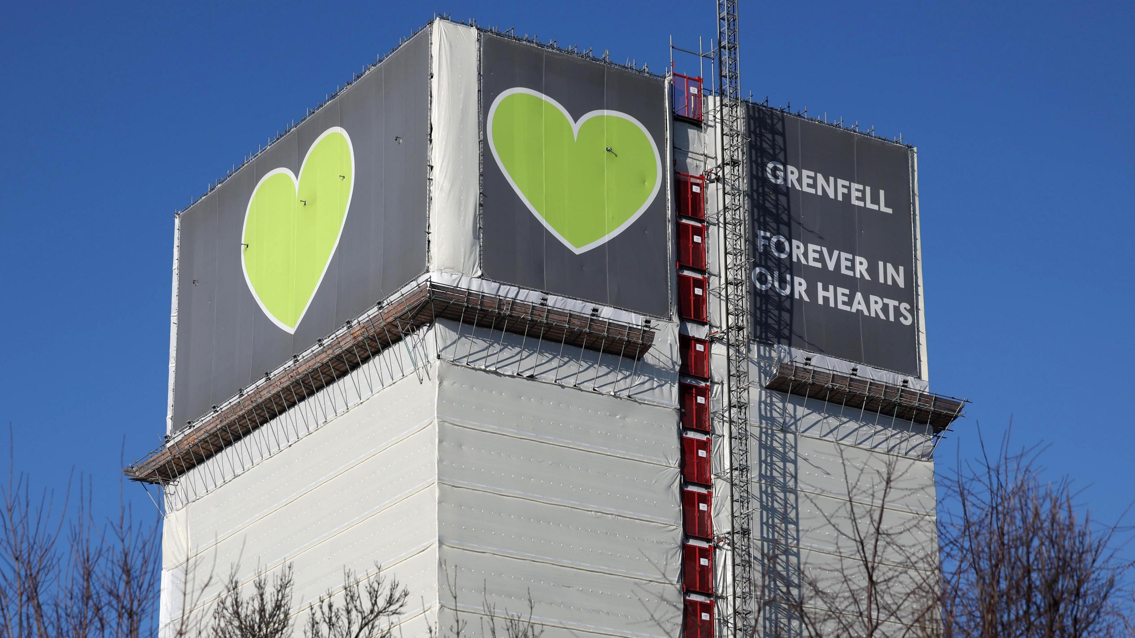 The top floors of Grenfell Tower in London, covered up with material featuring signs showing two large green hearts and the words 'Grenfell - forever in our hearts'