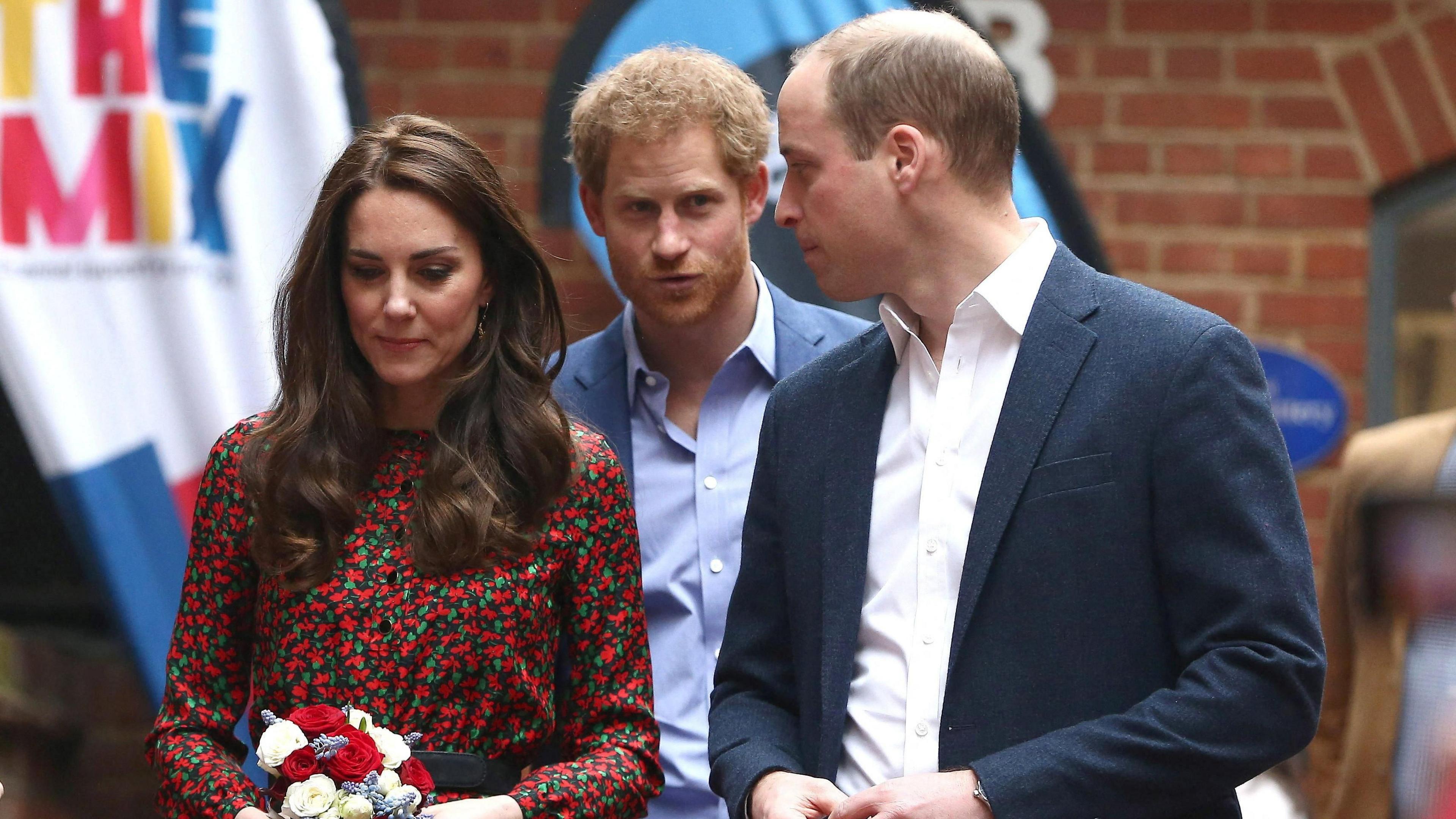 The Princess of Wales, dressed in a red floral dress, and Prince of Wales, in a blue suit, stand in front of the Duke of Sussex