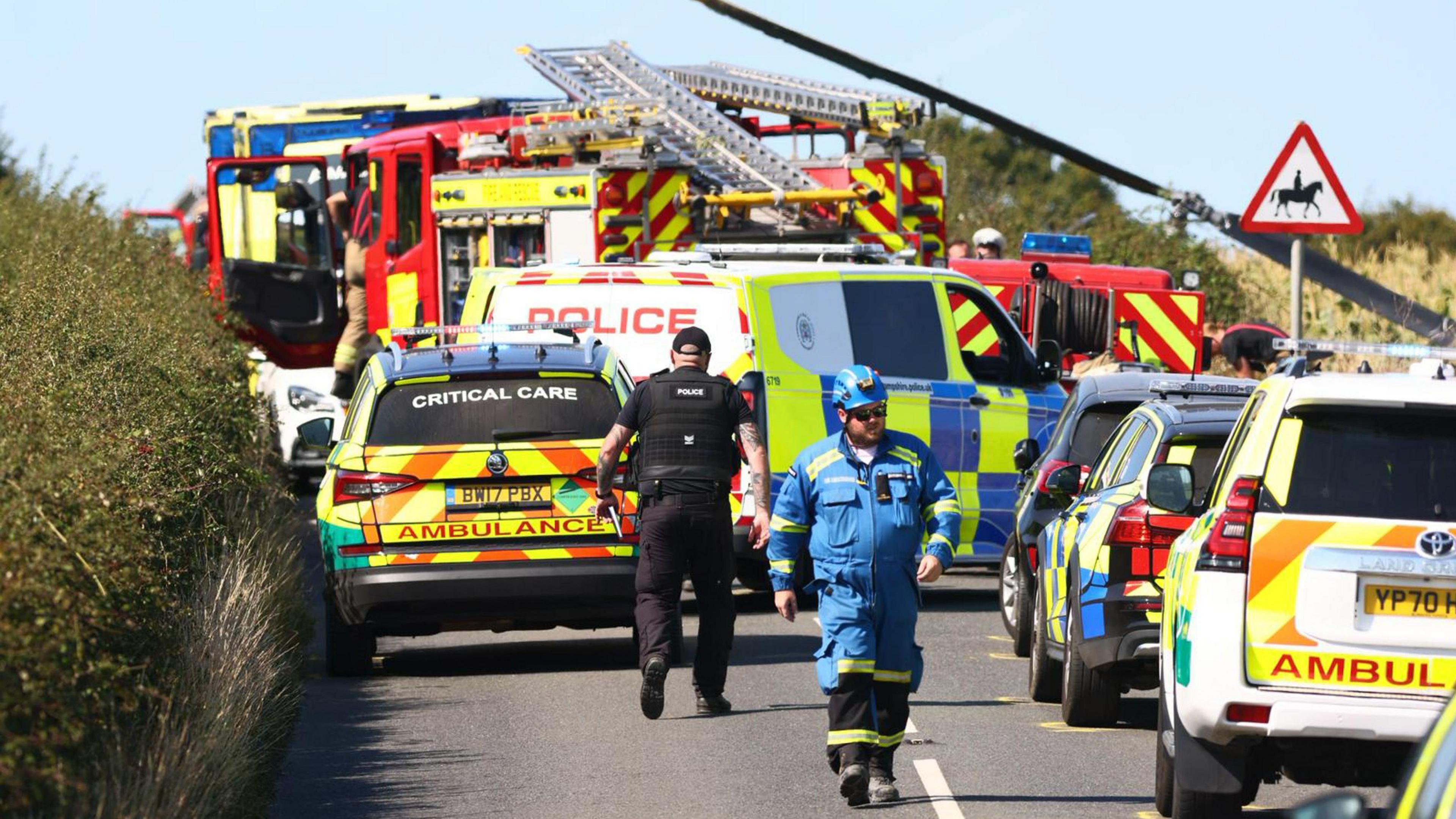 Emergency services including police officers and a man in all blue overalls, walking on a road with emergency service vehicles parked along it