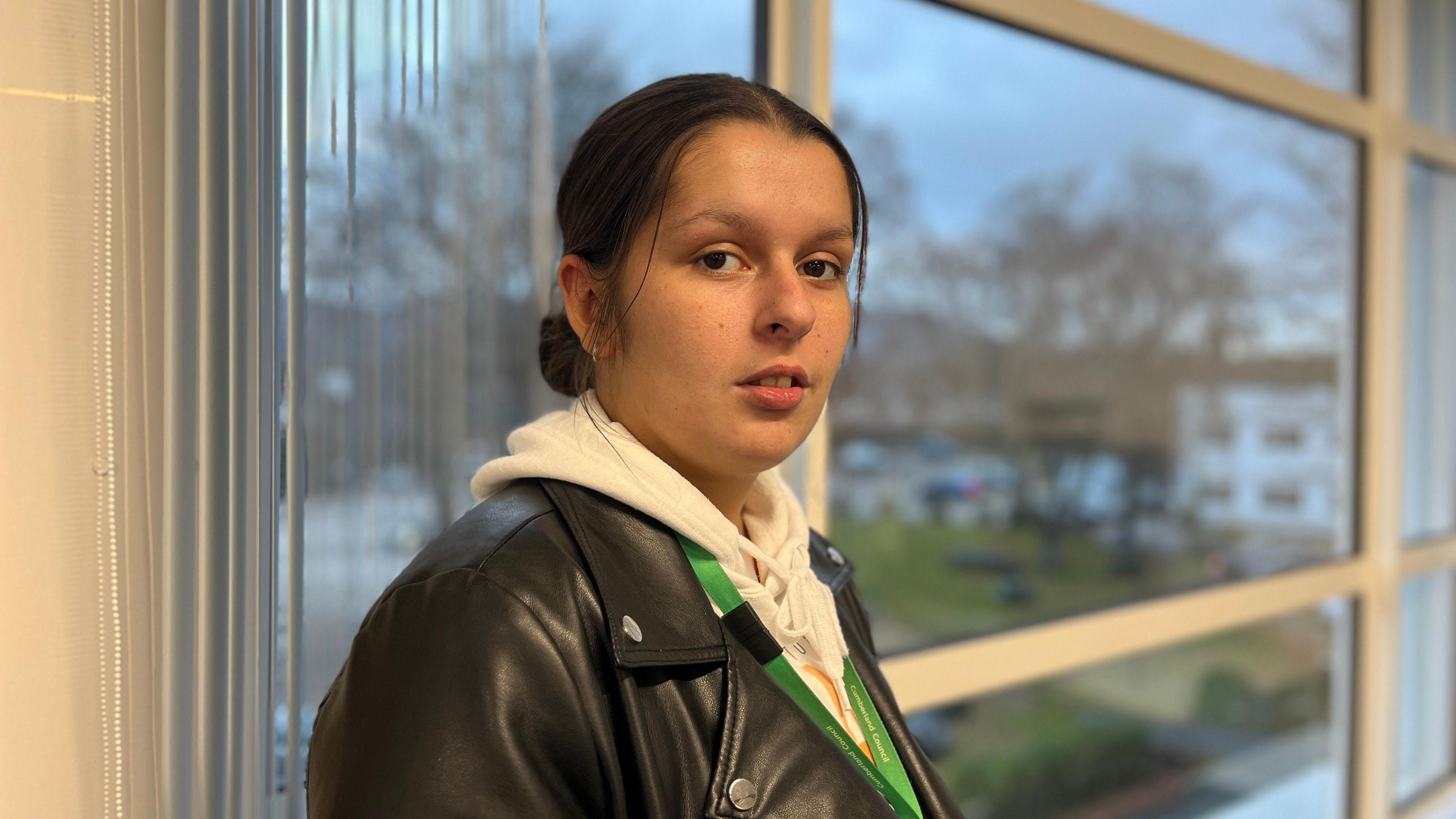 Chloe Morley standing in from of a window at the Keswick Pencil Factory. She is looking at the camera with a serious expression. She had long dark hair in a ponytail, dark eyes and is wearing a black leather jacket on top of a white hoodie.