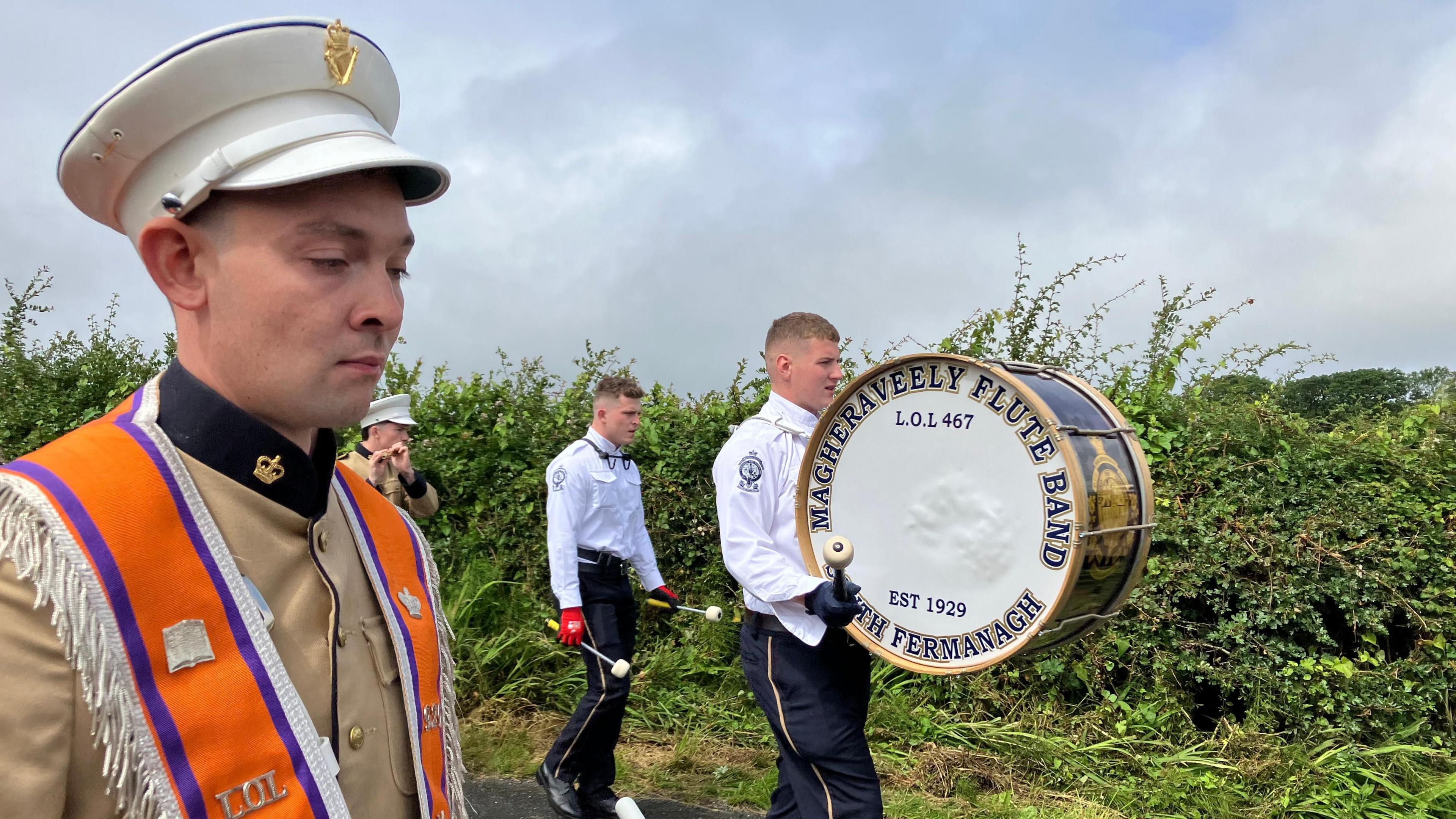 Three men marching. The man closest to the camera is wearing a white hat, brown blazer, and an orange sash around his shoulders. To his right is a man with a large drum which says 'Magheraveely Flute Band South Armagh'. 