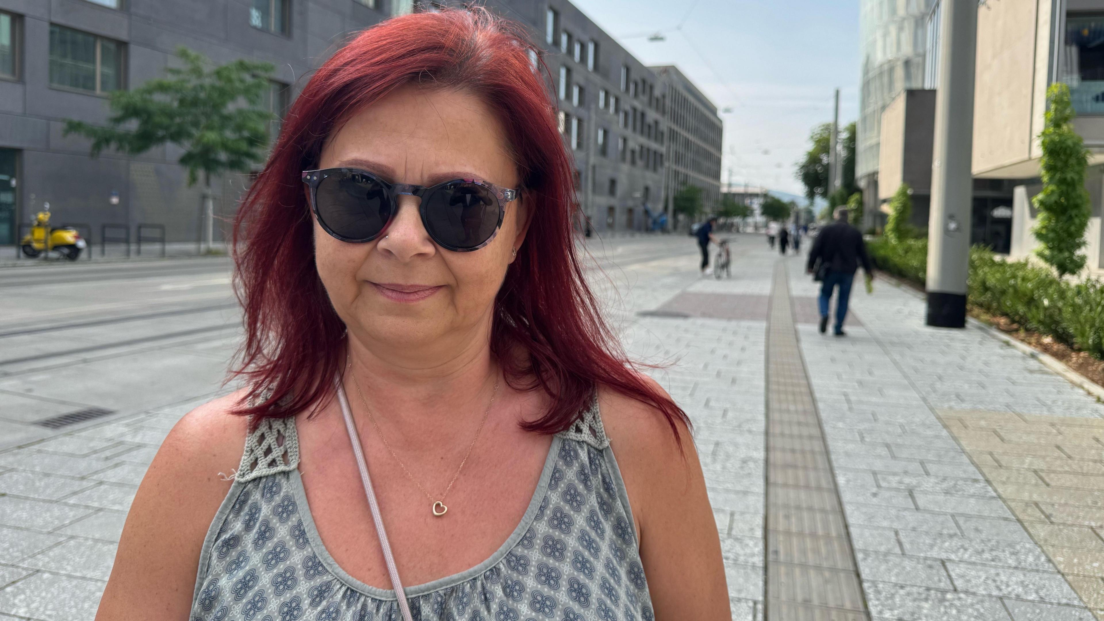 A woman with red hair and sunglasses looks at the camera