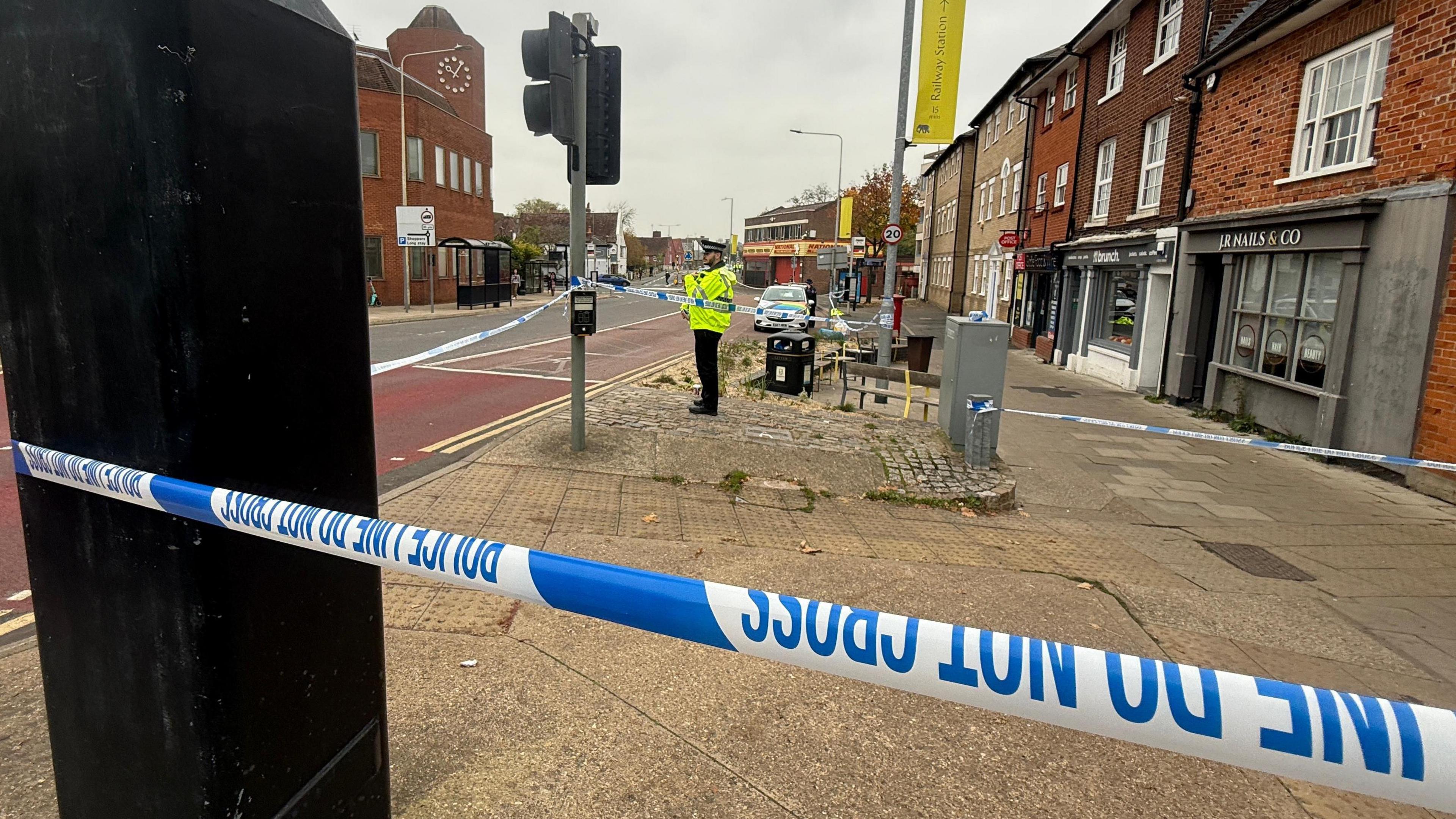 A police officer stands in front of an area cordoned off with blue and white police tape. It is all based on a pavement next to a road with shops. It is a grey, cloudy day.