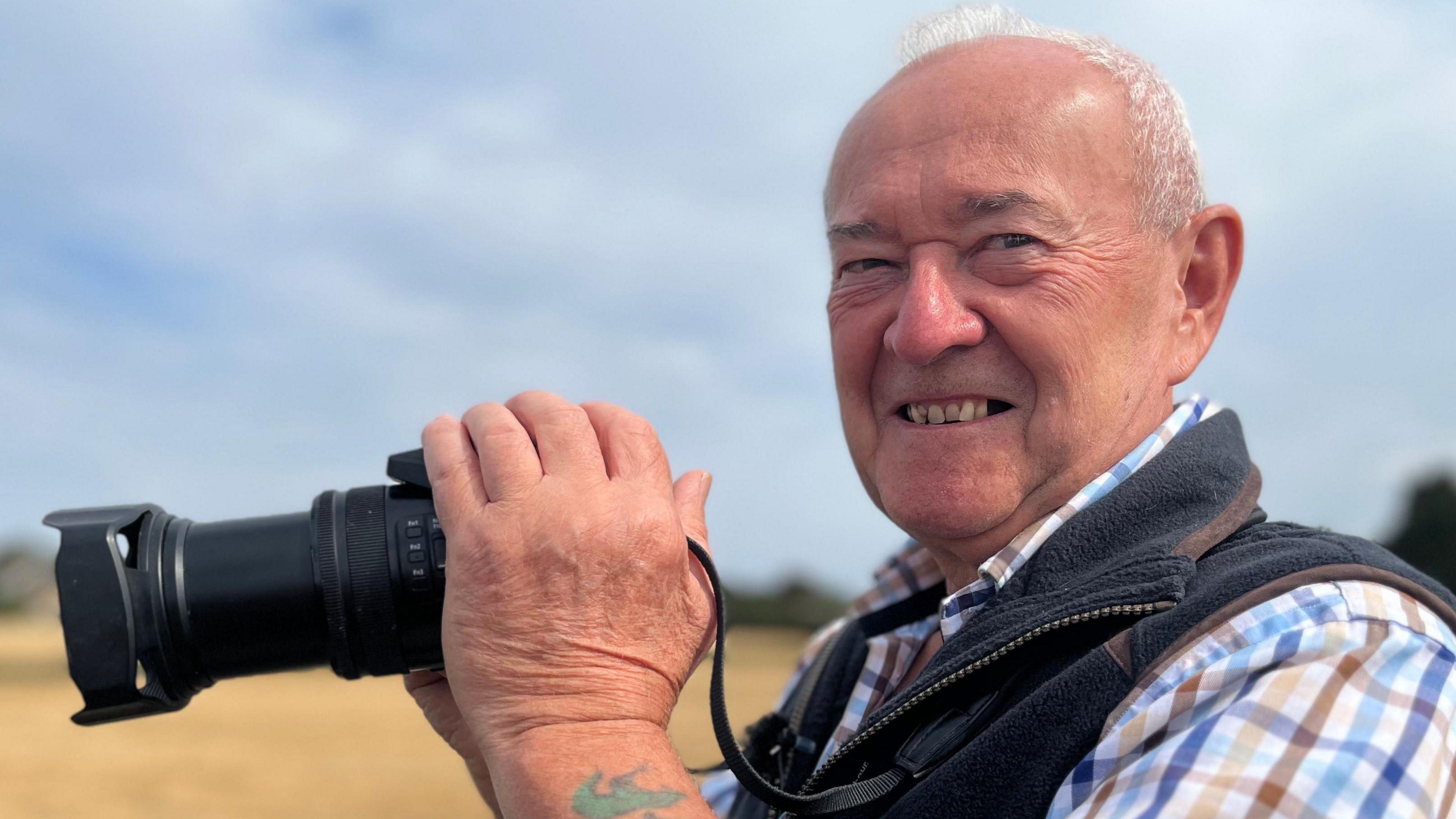 Dave Bradley is on the right side of the image looking towards the camera smiling. He is wearing a checked shirt and gilet and is holding a black camera.