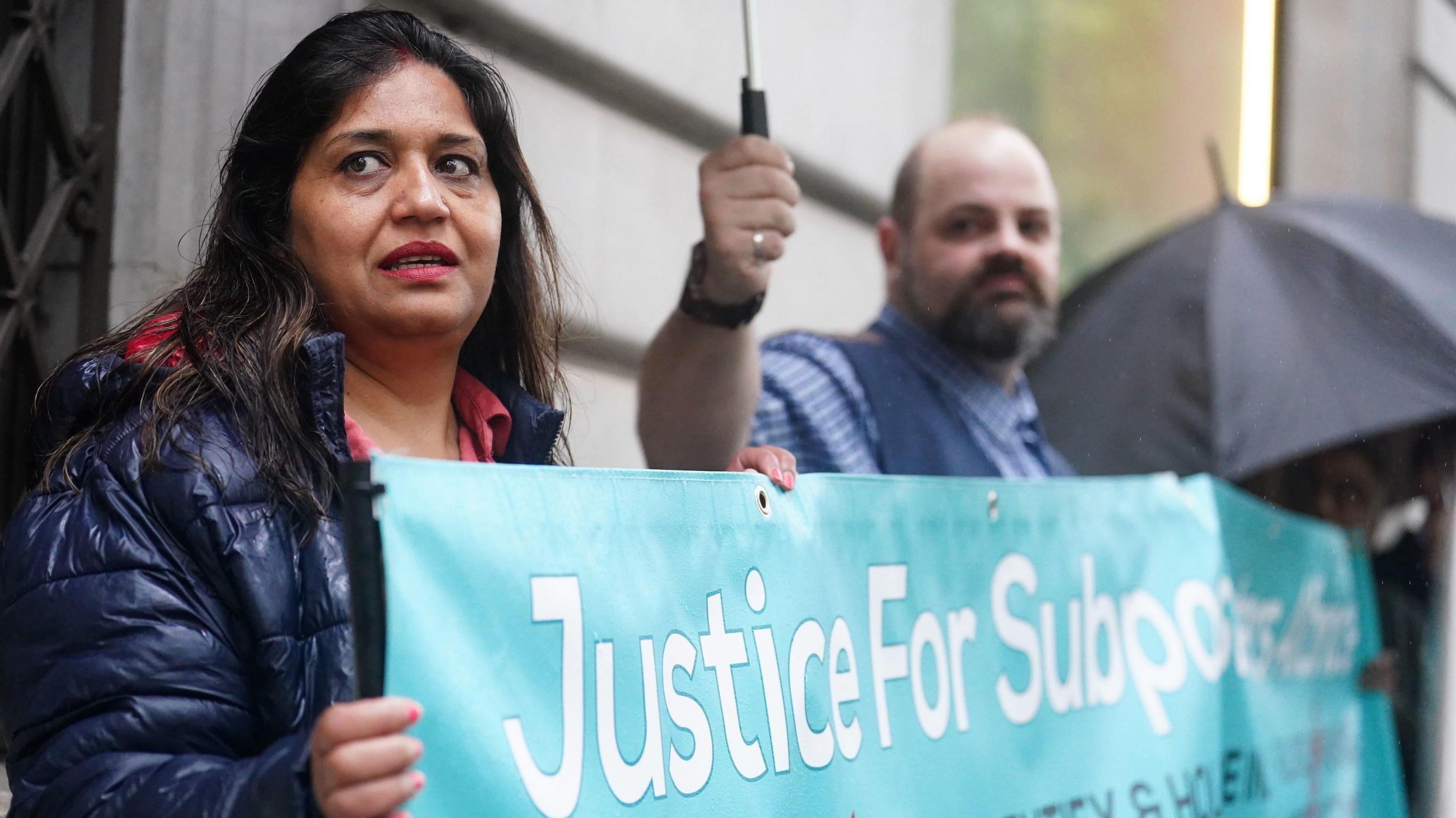 A woman with long dark hair wearing a purple jacket holds a banner saying Justice for Sub-postmasters Alliance