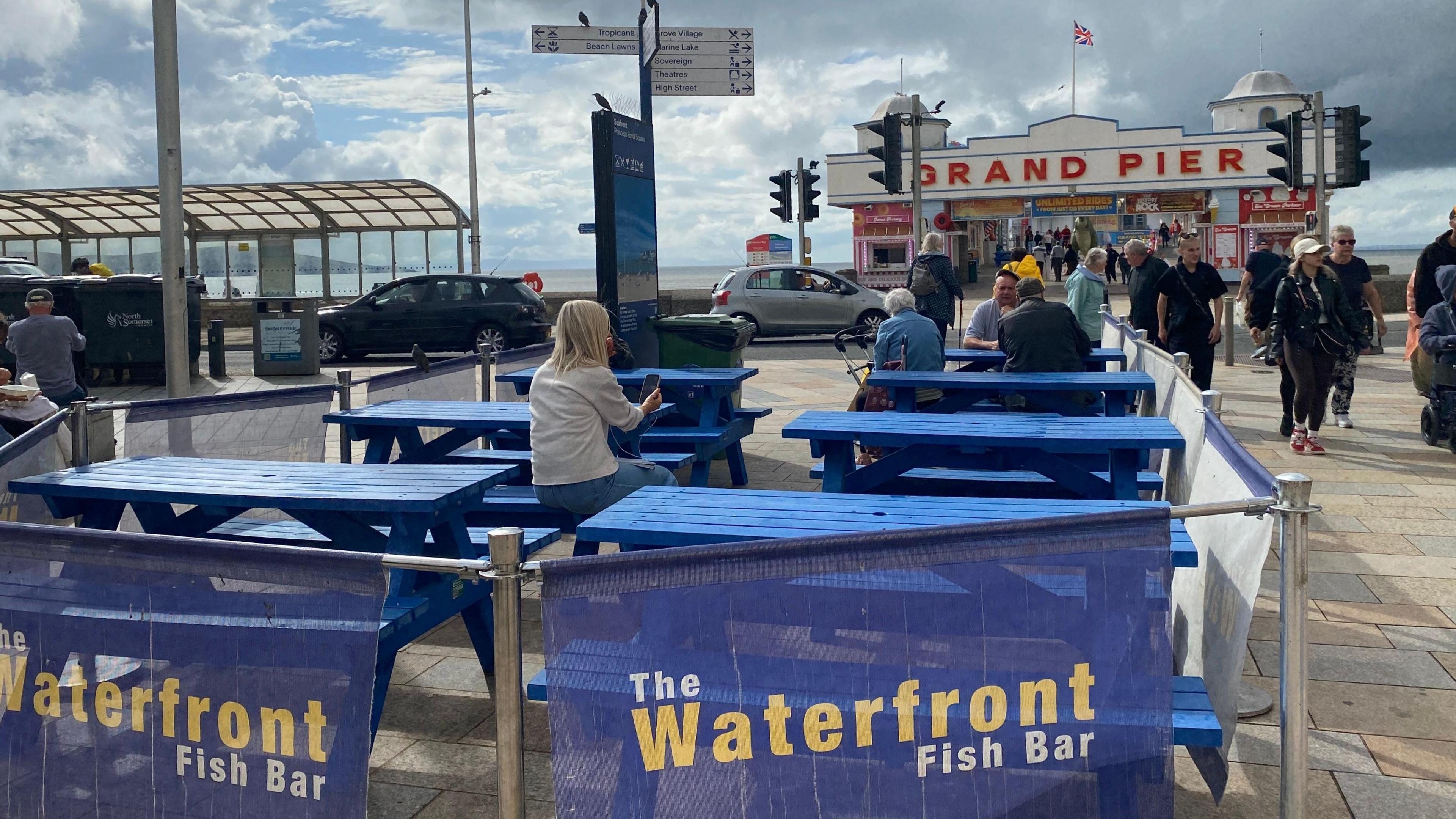Blue picnic tables are facing the water and the Grand Pier in Weston-super-Mare. Some people are sitting at the tables and walking past.