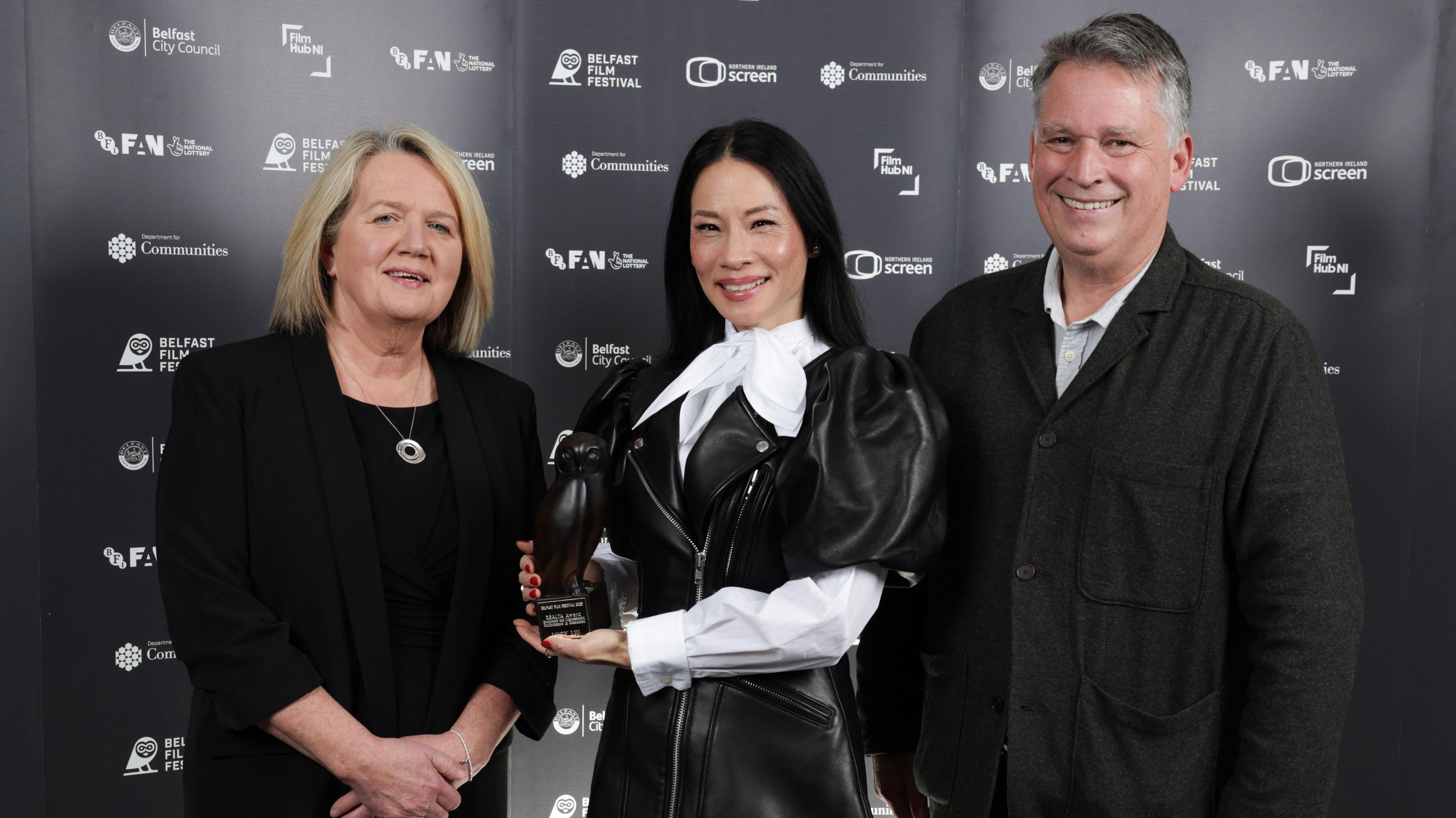 Lucy Liu poses for a photo while standing between Belfast Film Festival director Michele Devlin and Richard Williams from Northern Ireland Screen. Ms Devlin has shoulder-length blonde hair and is wearing a black suit and top. Mr Williams has short, greying hair and is wearing a dark jacket over a pale-coloured open-neck shirt. 