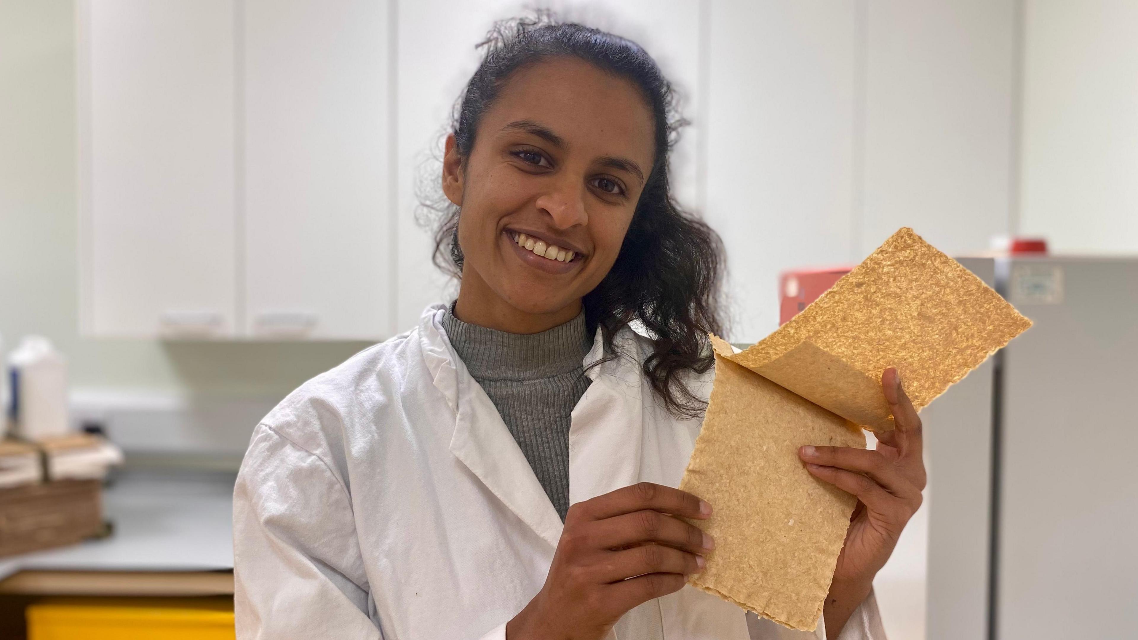 Renuka Ramanujam in a white lab coat holding some of the onion peel paper she has made. There are blinds, a bench and a filing cabinet behind her.