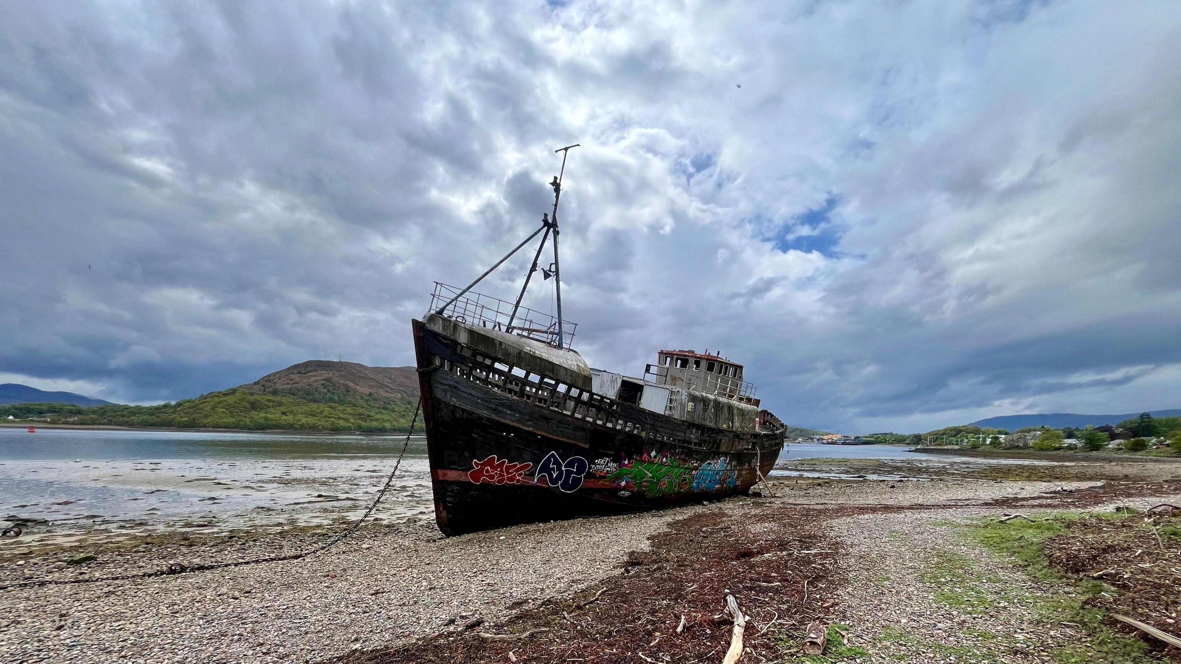 A graffiti-covered and possibly abandoned fishing boat beached on a shingle shore next to a lake or sea inlet, under a cloudy sky