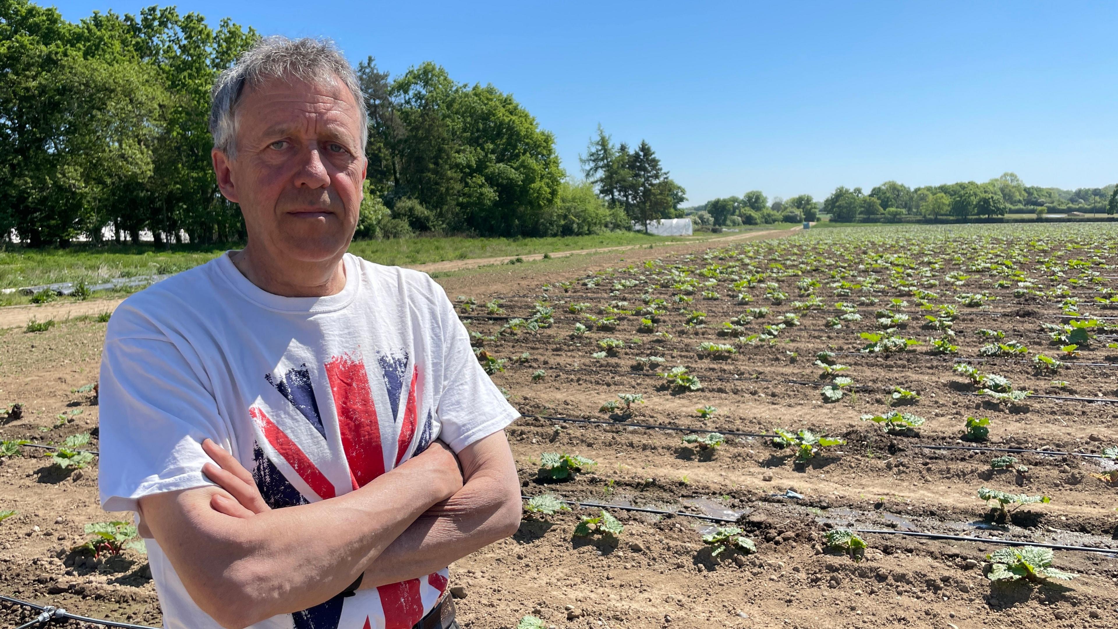A man stands with his arms crossed wearing a white t-shirt with a union jack on it. Behind him is a dry-looking field with some small plants in rows in the soil.