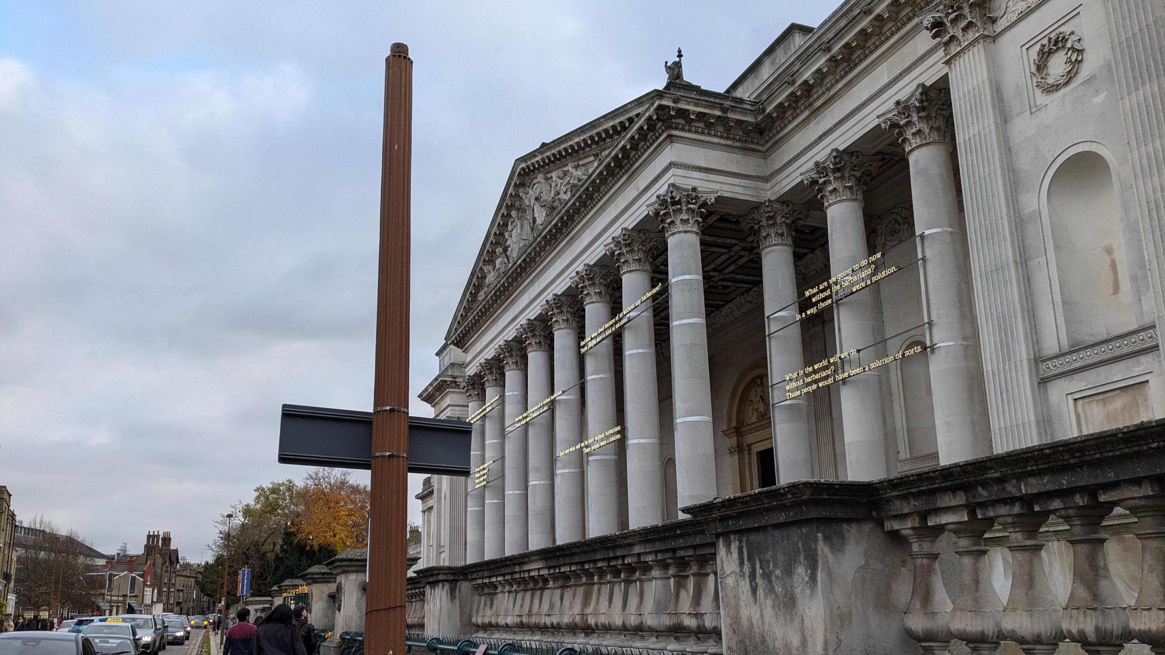 The front of the Fitzwilliam Museum has ornate columns outside. On the path outside is a post which is missing the lamp on top. There are people walking on the pavement and cars driving down the road