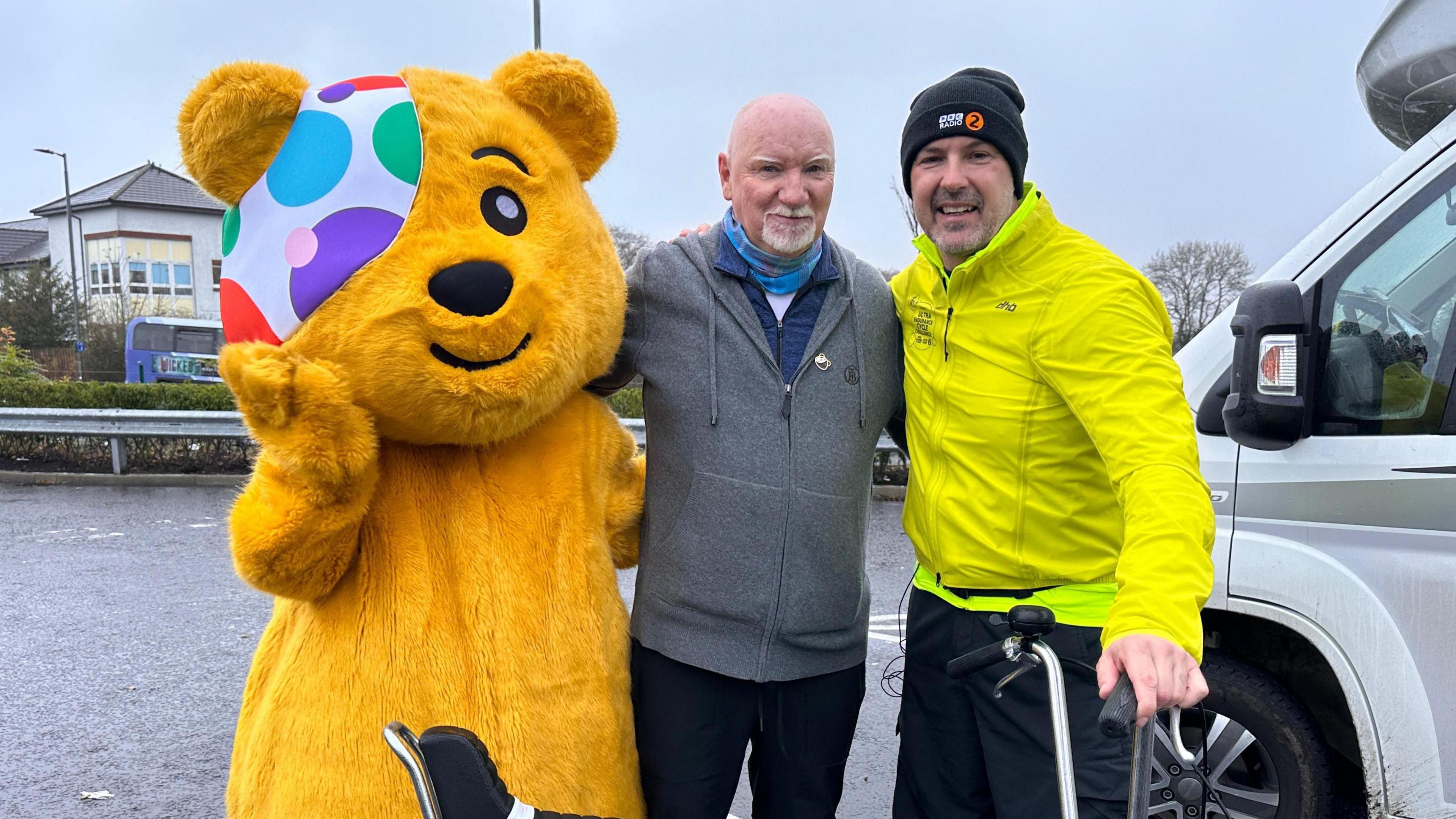 Pudsey, Sir Tom Hunter and Paddy McGuinness with arms around one another, standing next to a chopper