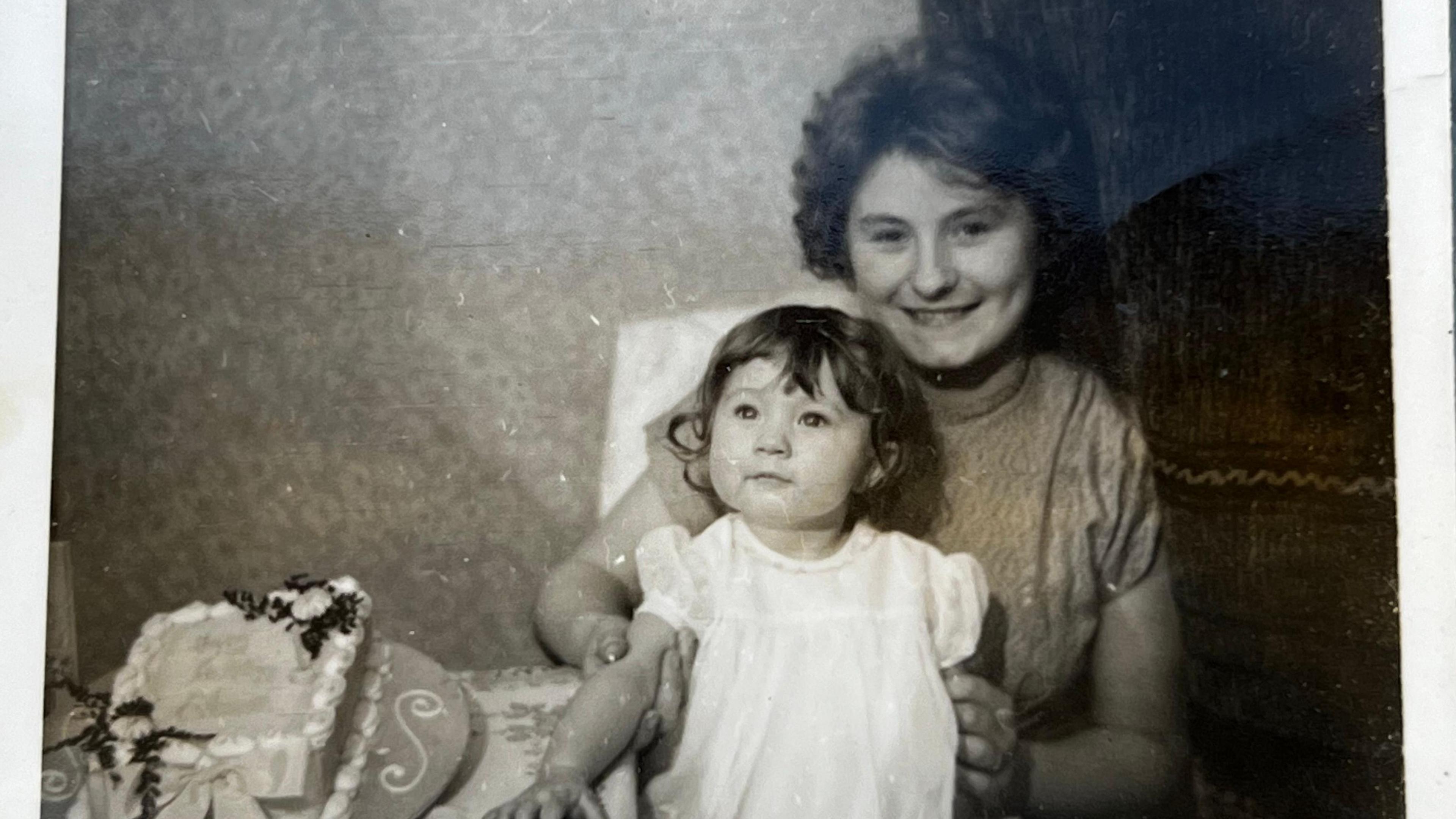 A black and white photo of two young girls, one of whom is significantly younger than the other. The older girl holds the younger girl's arms and smiles at the camera.