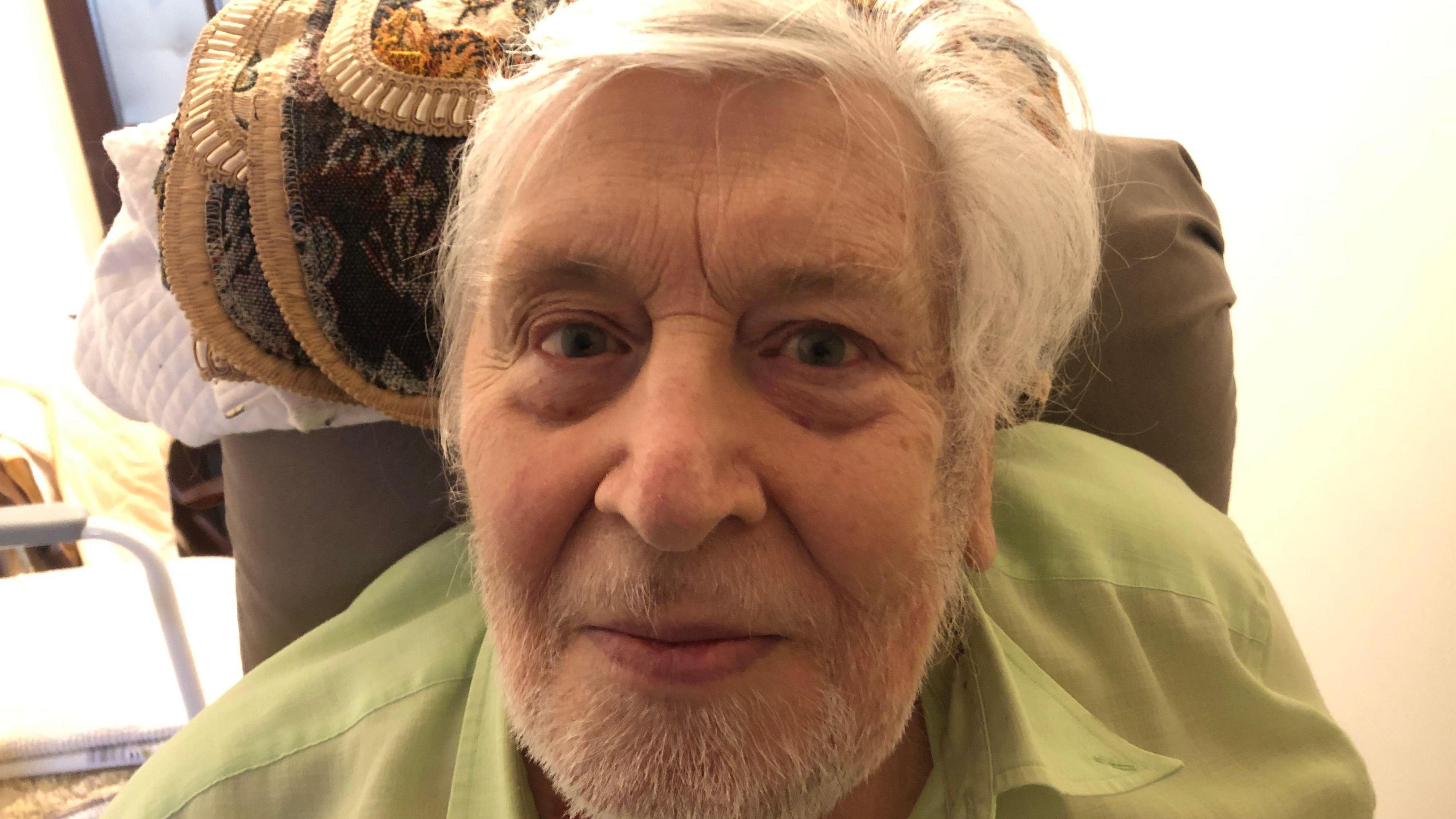 Peter Deadman, 84, has white hair and a white/grey beard and moustache. He is wearing a light green shirt and is sitting in a high-backed chair