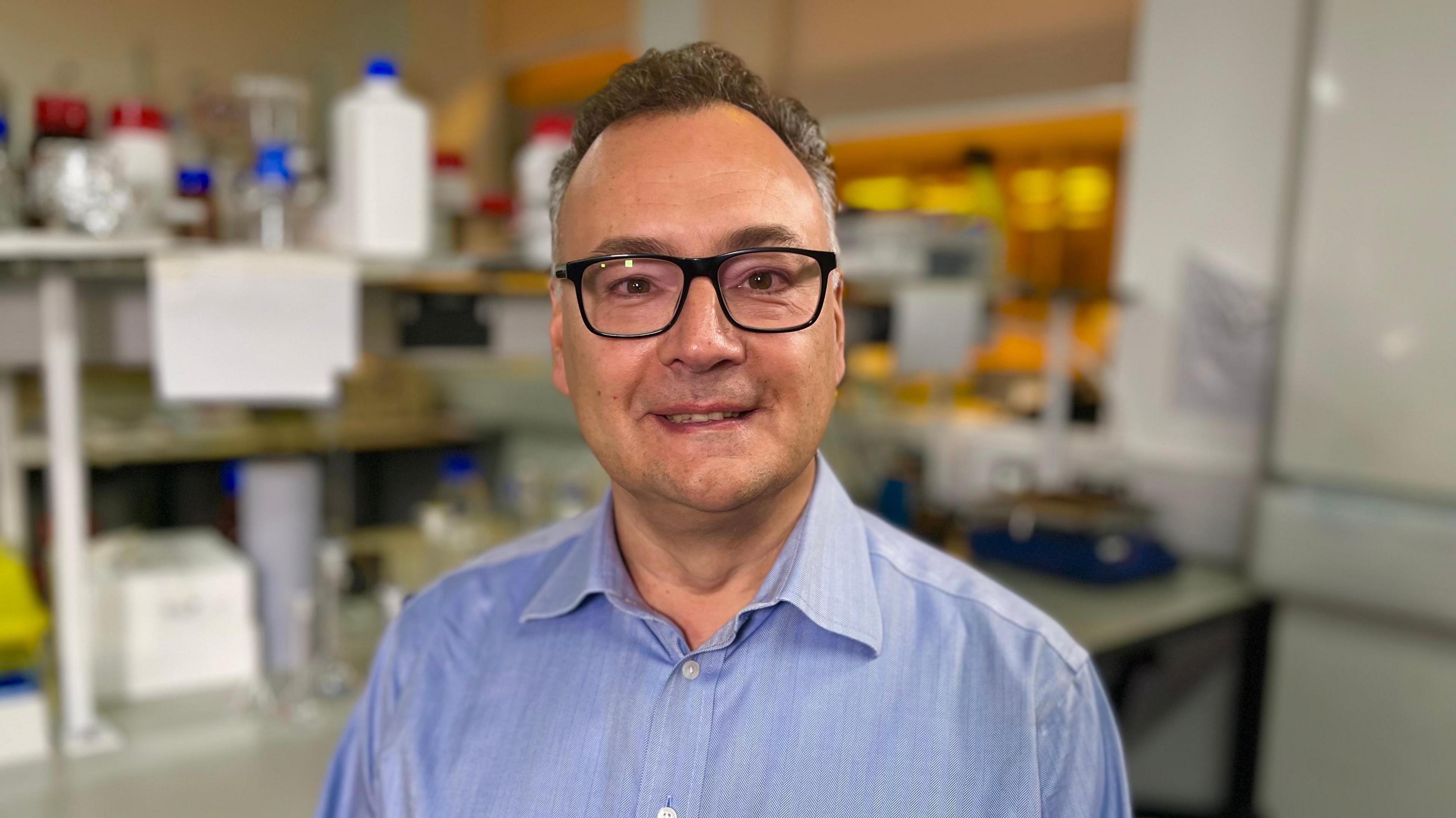 Professor George Malliaras in a lab. He has short grey hair, dark framed glasses and is wearing a lilac shirt. He is smiling into the camera.