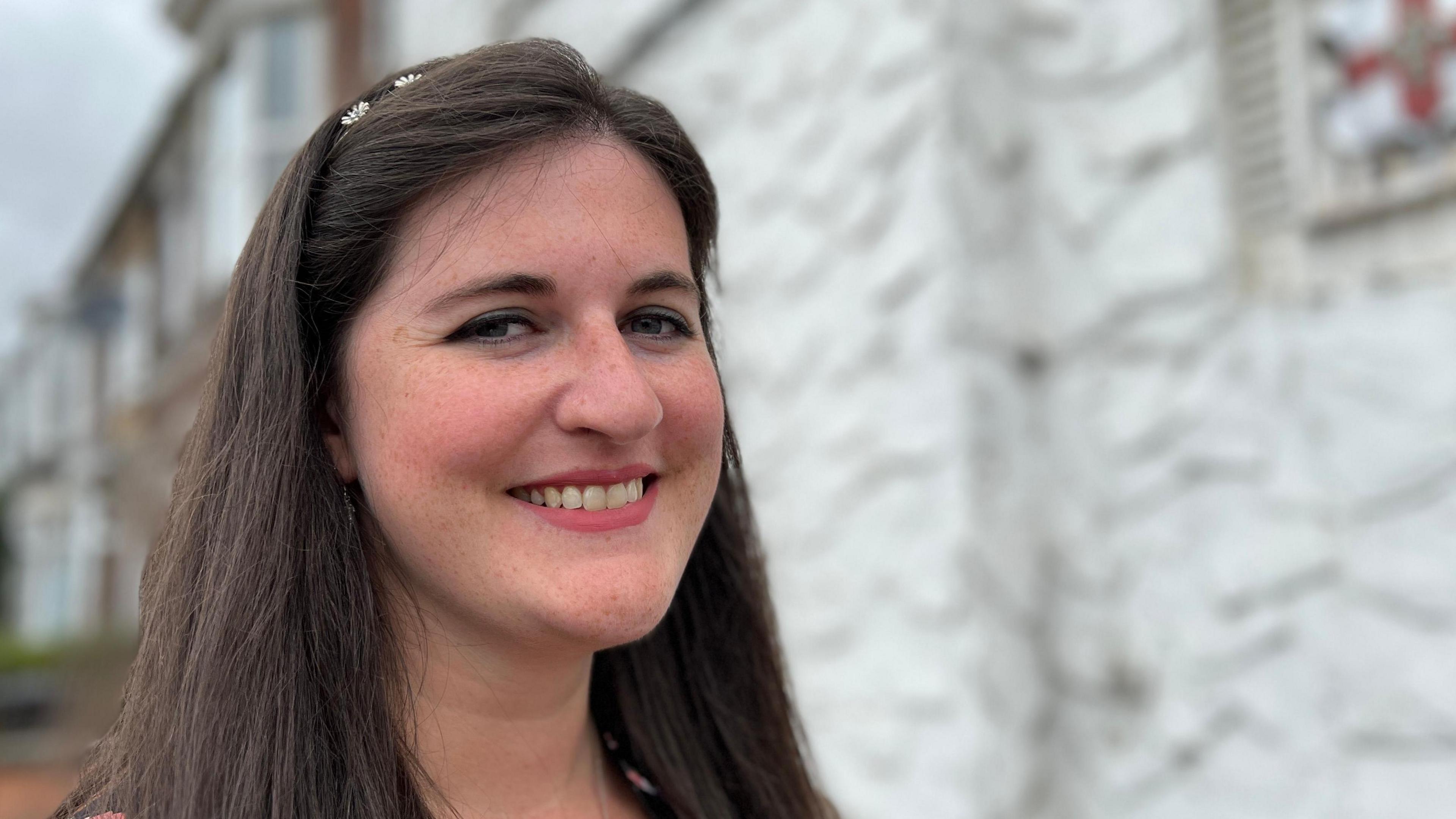 Headshot of Eleanor Bryan with long brown hair smiling with teeth into the camera in front of the house